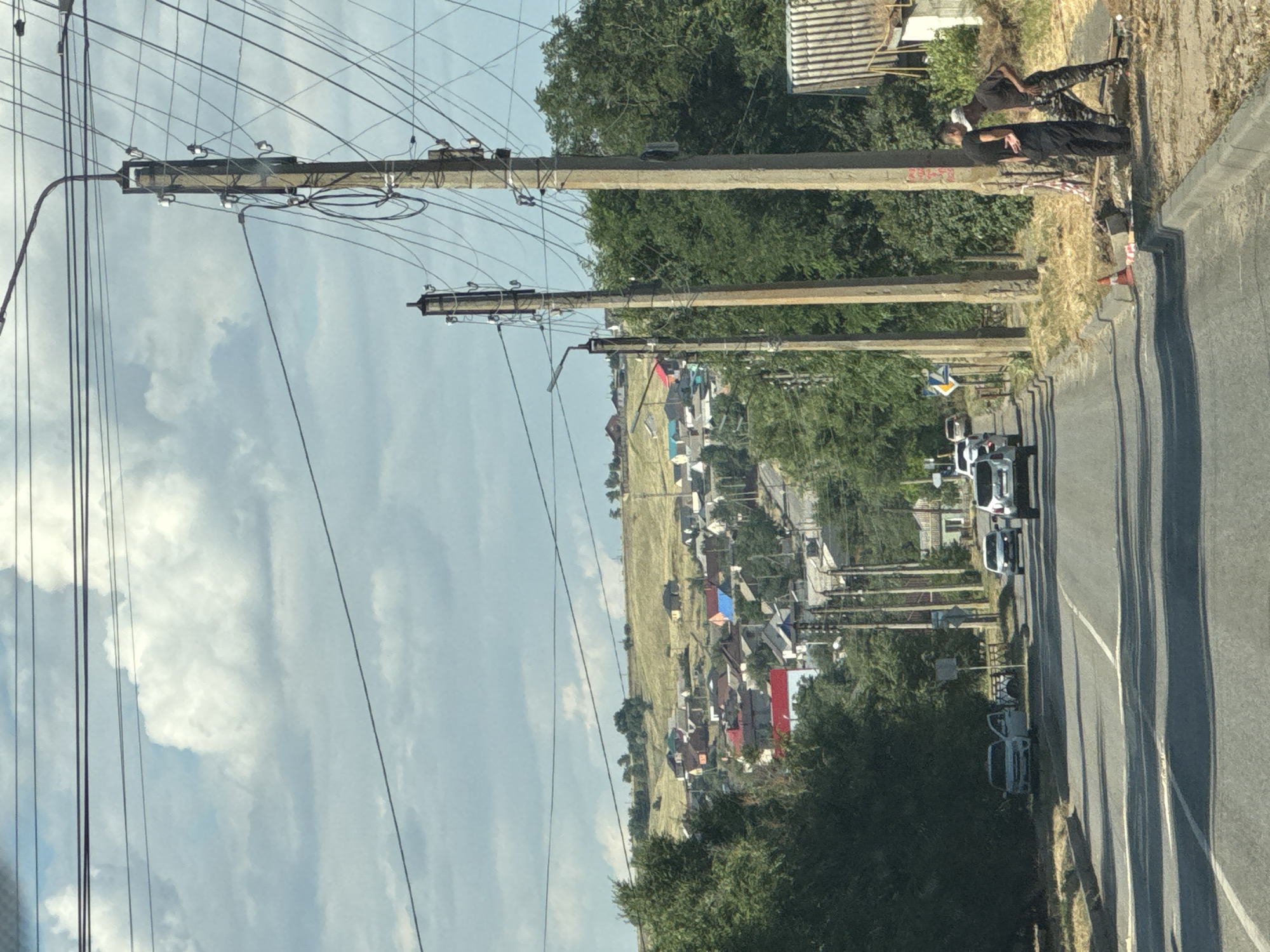The image depicts a roadside scene in a rural area, likely in Russia given the location context. The orientation of the image is rotated, showing a street on the right side and a field with houses on the left, separated by a line of utility poles and trees. The sky is partly cloudy, suggesting daytime with fair weather.
In the foreground on the right side of the road, two individuals are visible. One person is standing, leaning against a structure, and another is seated. They appear to be engaged in some casual activity or waiting. Further down the road, several cars are driving or parked. One vehicle, a white SUV, is prominent. On the left side of the road, a field is dotted with houses, many with colorful roofs, indicating a residential area within a village or small town. The utility poles are a prominent feature, carrying numerous wires that crisscross the frame, suggesting a well-connected area despite its rural setting.
Notable details include the presence of traffic cones and construction barriers near the road, hinting at ongoing work or road maintenance. The overall impression is one of a normal day in a rural Russian settlement, with daily activities and infrastructure visible. No specific text is clearly legible in the image.