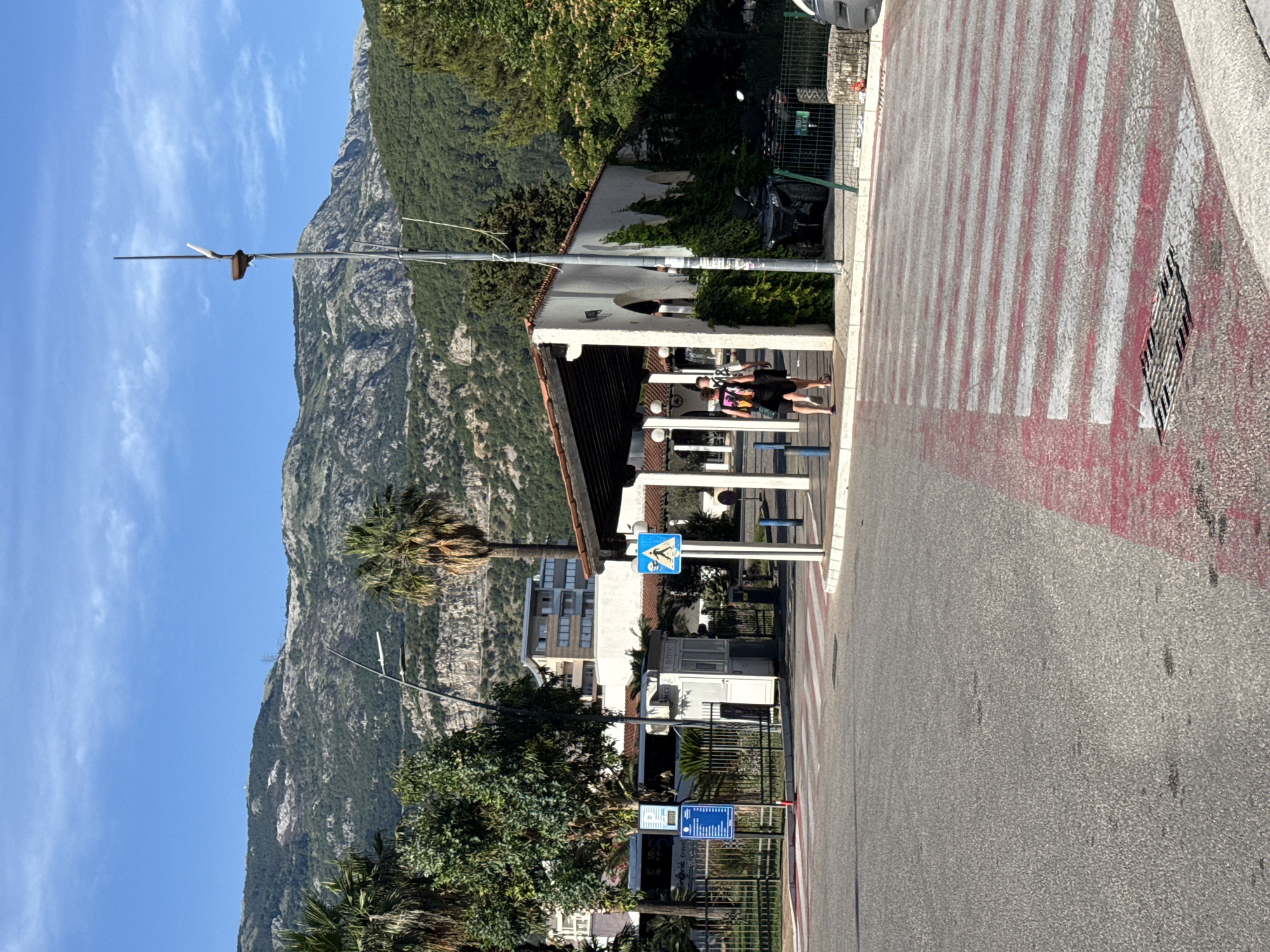 The image depicts a street scene in Budva, Montenegro, on a clear day. In the foreground, a red and white striped pedestrian crossing spans across asphalt. To the left, a traffic light pole stands, and beyond it, a white booth with a sign reading "ENTRANCE" is visible. Further left, a bus with "SINQVIC TRAVEL" written on it is parked behind a metal fence. Behind the booth and fence, a multi-story building with modern architecture is visible. To the right of the crossing, a covered walkway with white pillars and a tiled roof extends. Two individuals, one wearing a backpack, are walking away from the camera under the walkway. A street lamp with a peculiar design is situated to the right of the walkway. In the background, lush green trees and palm trees are interspersed with buildings, leading to a mountainous landscape under a bright blue sky. A parked all-terrain vehicle is visible behind the fence to the right of the walkway.