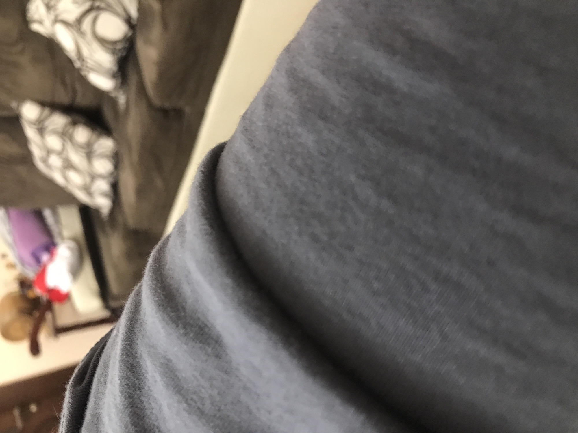 The image shows a close-up view of a gray fabric, likely clothing, in the foreground. In the background, there is a brown couch with patterned pillows. A small table with various items, including a purple object, a red and white object, and a brown object, is also visible. The location information is not discernible from the image.