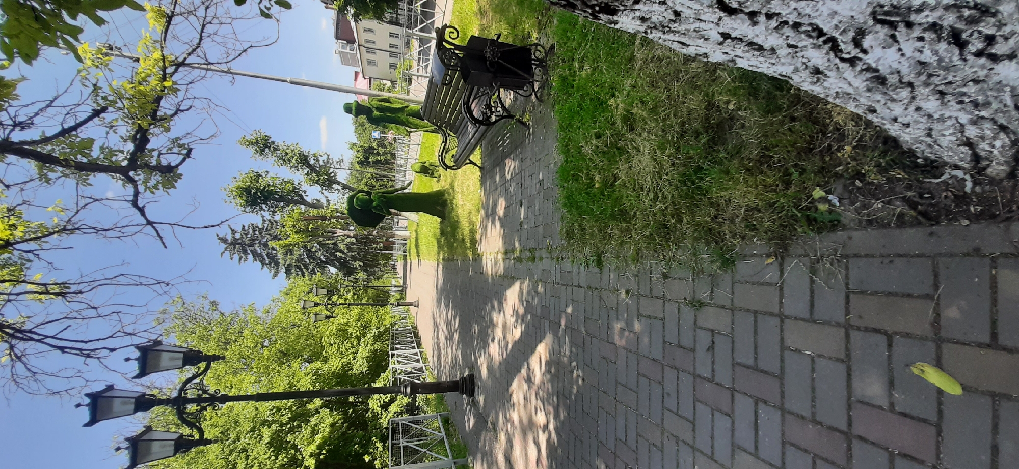A paved walkway lined with trees and a decorative fence runs through a park. A three-lamp street light stands to the left of the walkway. Further down the path, a park bench with a trash receptacle sits near a topiary shaped like a woman holding an umbrella and another topiary resembling a man in a hat, with a smaller topiary of a dog between them. A single yellow banana peel lies on the walkway in the foreground. The sky is clear and blue, and sunlight casts shadows across the path. Buildings can be seen in the distance on the right side of the image. The scene depicts a public park in Yessentukskaya, Russia, on a sunny day.