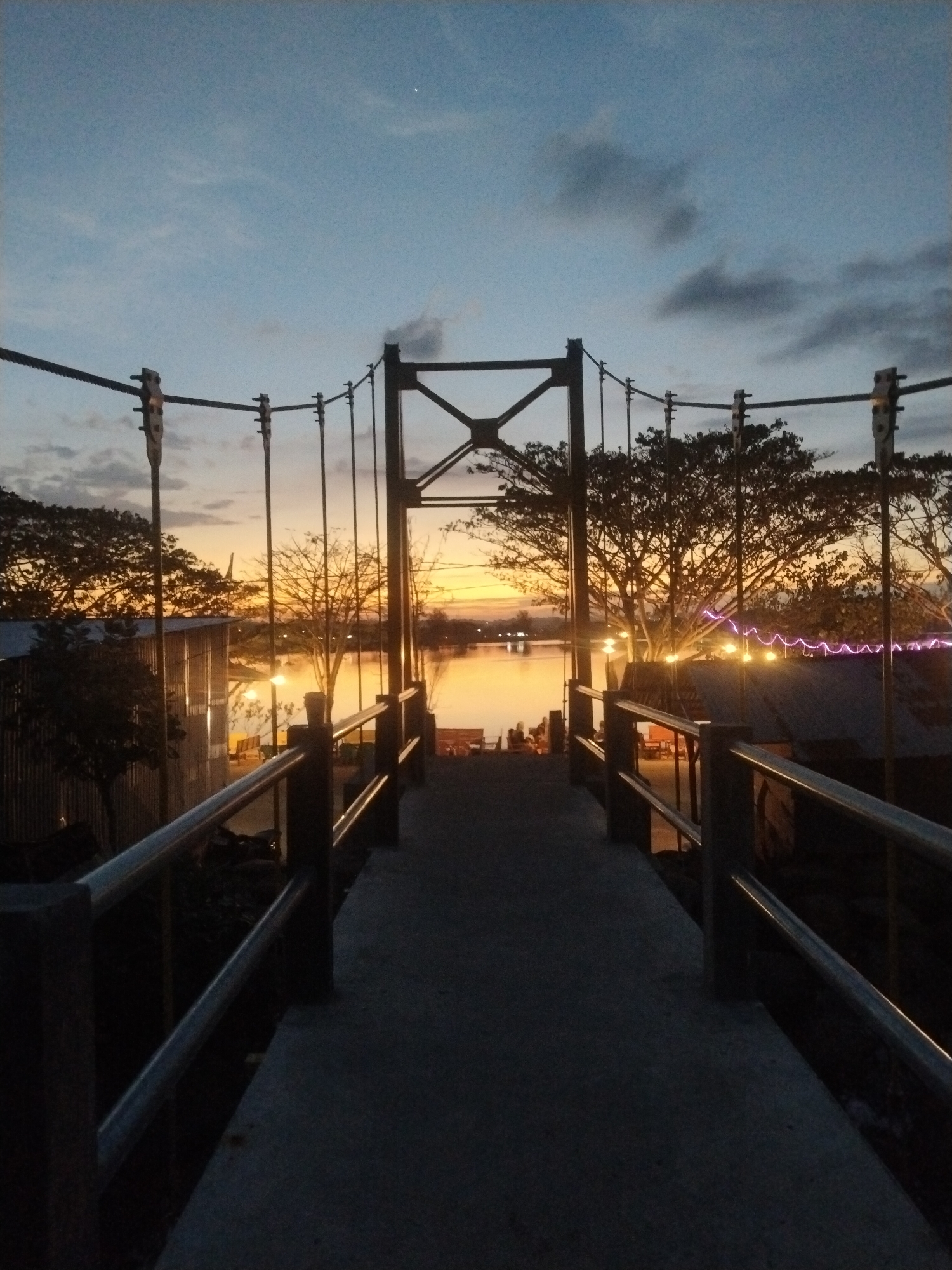 The image shows a pedestrian suspension bridge in Lhokseumawe, Indonesia, at twilight. The bridge deck is made of concrete with metal railings. Suspension cables run above, attached to metal support towers. Beyond the bridge, a body of water reflects the orange and yellow colors of the setting sun. Buildings and trees are visible on the opposite shore, illuminated by artificial lights. The sky is a gradient of orange, yellow, blue, and gray. Small groups of people are visible on the far side of the bridge. A single bright star is visible in the darkening sky.