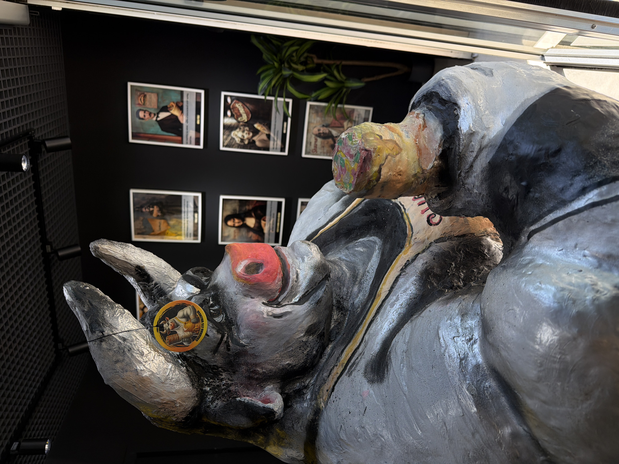 A close-up shot depicts a painted sculpture of a rhino head, predominantly grey and black, with a red nostril. A circular tag hangs from one of the rhino's horns, featuring a caricature of a rhino wearing a yellow outfit, possibly a reference to Valencia's "Falles" festival.
In the background, dimly lit, are several framed pictures on a dark wall. To the left is a grid-like wall with black lights. The scene appears to be indoors, possibly in a gallery or exhibit. Based on the tag and the style of the artwork, this scene is likely located in Valencia, Spain.