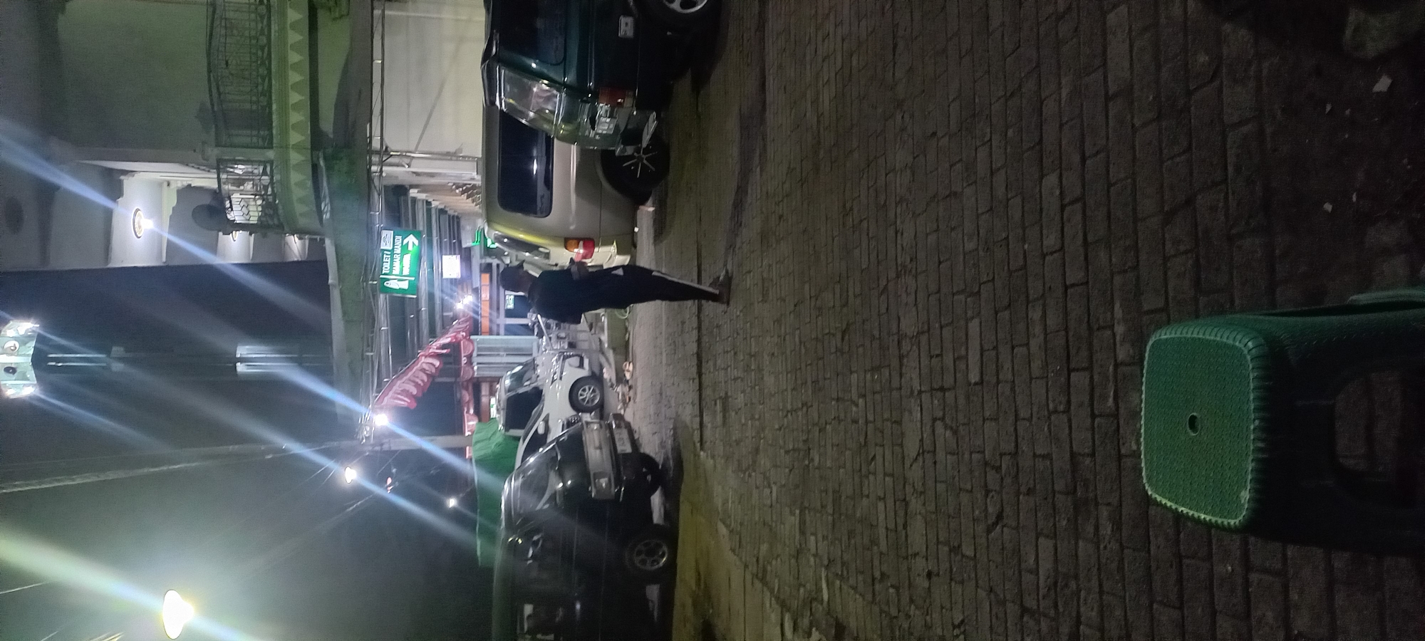 The image depicts a nighttime scene in what appears to be an outdoor area, possibly a parking lot or walkway, paved with brick-like stones. In the foreground, a green plastic stool is visible. Further back, a person wearing a dark shirt, dark pants with white stripes, and a dark cap stands facing away from the camera, looking at their phone. They are positioned between several parked vehicles, including a black van, a white van, a gold minivan, and a dark green SUV. The background features elements of a building, including a tower with illuminated sections, a balcony with railings, and a green sign with white text that reads "TOILET / KAMAR MANDI" and an arrow pointing to the right. Overhead lighting casts strong beams of light across the scene, creating glare. The presence of the "TOILET / KAMAR MANDI" sign, combined with the architecture and vehicles, suggests the location could be a public facility, such as a rest area or a place of worship with amenities, in Banjarsari, Indonesia.