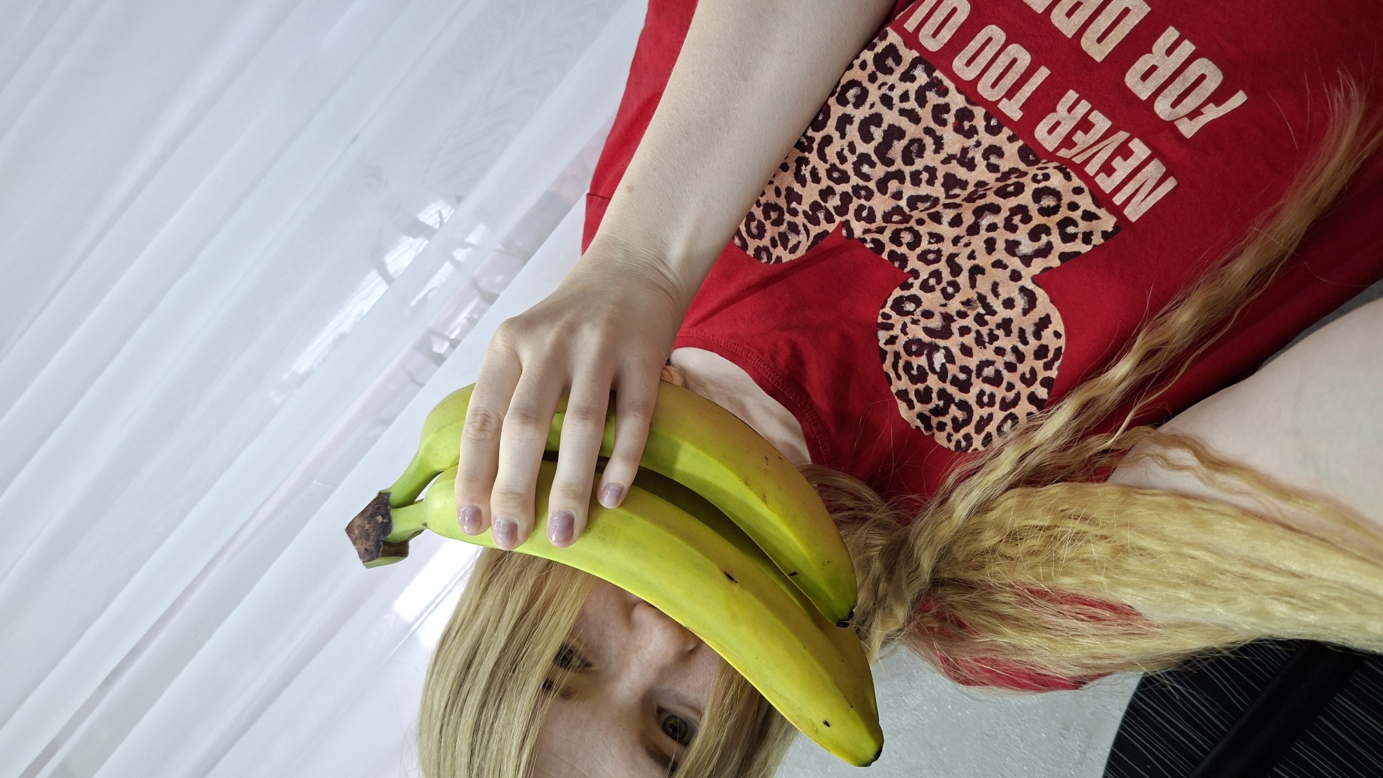 This rotated close-up image features a person, likely a woman, lying down and holding a bunch of bananas. The subject's face is partially obscured by the bright yellow-green bananas, but one eye, framed by blonde hair and bangs, looks directly at the viewer with a neutral expression. Their hand, visible holding the bananas, has light-colored nail polish.
The person is wearing a red t-shirt with a large leopard print design in brown and black. Visible text on the shirt, printed in a lighter color and appearing upside down relative to the subject's orientation, reads "NEVER TOO OLD DR...". The blonde hair is long and slightly wavy, cascading around the person's head and shoulder.
The background is softly lit by ambient light filtering through sheer, white curtain-like fabric, suggesting an indoor setting, possibly near a window, during the daytime. The bananas appear fresh, with some green remaining, indicating they are not fully ripe. This intimate portrait-style shot is set in Chaikovsky, Russia.