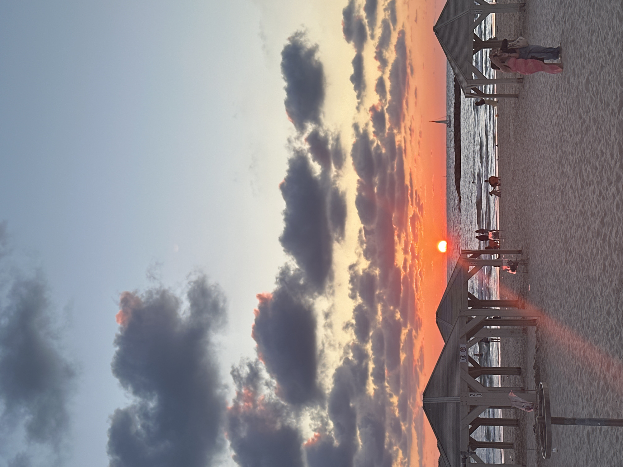 Captured at sunset on a beach in Tel-Aviv, Israel, the image presents a blend of natural beauty and human presence. The sky is a dramatic mix of light blue meeting a vibrant orange sky, dominated by textured clouds tinged with shades of gray and orange. Below, the sun sets over the sea, casting a warm, intense glow. Wooden beach canopies line the sandy shore. A small sailboat is seen far on the horizon. People are present on the beach, either standing beneath the canopies or closer to the water. The sand is light-colored.