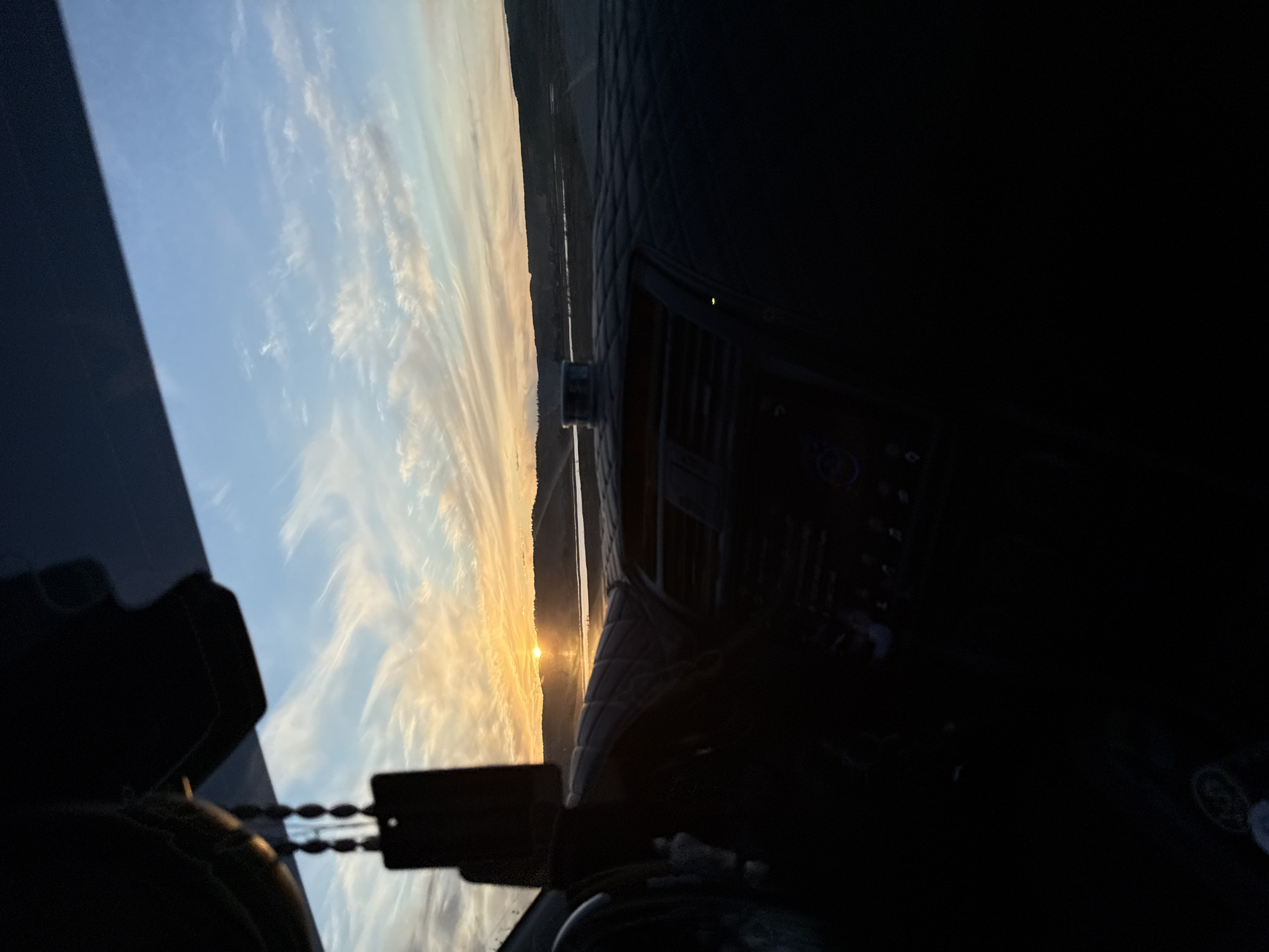 The image depicts a view from inside a vehicle, likely a car, facing a sunset. Outside, the sky is visible with a blend of blue and orange hues. Wispy clouds stretch across the sky. A body of water or flat terrain stretches horizontally across the middle distance. The dashboard of the vehicle is visible in the foreground, partially obscuring the view. The location is uncertain; there is no identifiable evidence to suggest it is Kil'demtsy, Russia.
