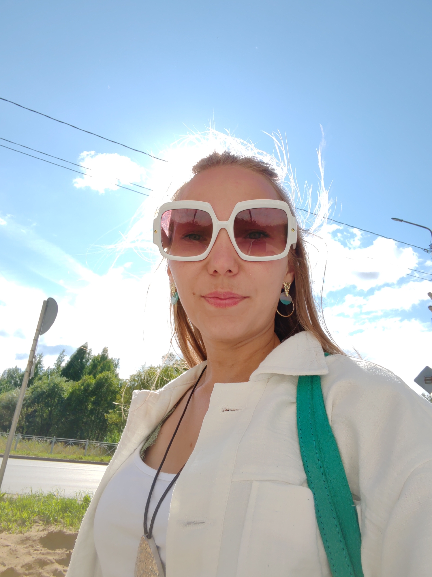 A woman stands outdoors on a sunny day, with the bright sun creating a lens flare and blowing her hair. She is wearing large, white-framed sunglasses with pink lenses, a white collared shirt over a white tank top, and a distinctive necklace with a large, flat, pendant. She also has on unique earrings with a gold triangle and blue and teal circular elements. A bright green strap from a bag is visible across her shoulder. The background shows a blue sky with some clouds, telephone wires, trees, a road, and a traffic sign. The overall impression is of a casual, stylish portrait captured in an urban or suburban outdoor setting. The image lacks specific visual cues that would definitively place it in Syktyvkar, Russia; however, given the provided context that it is from that location, it is assumed to be a scene within the city.