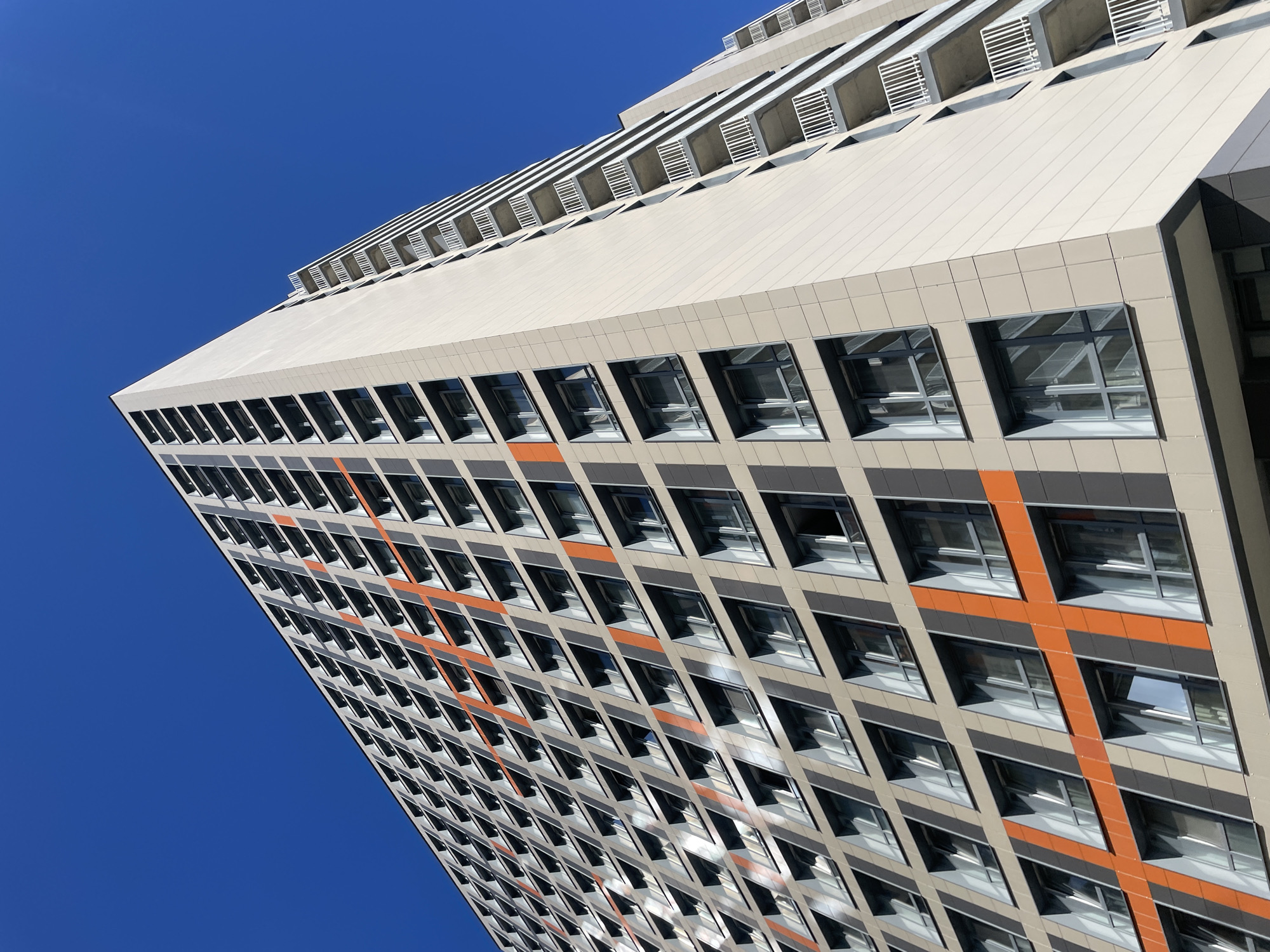 This is a low-angle, full shot of a modern, tall apartment building against a clear, bright blue sky. The building is constructed with light beige and dark grey panels, and features a striking pattern of orange diagonal lines across its facade, creating a visual grid. Numerous windows are visible, many with balconies that have white railings. The perspective emphasizes the height and scale of the structure, with its geometric design. The scene is devoid of people or any visible activity, presenting a clean, architectural view. The building appears to be located in the city of Saint Petersburg, Russia, given the architectural style and urban setting suggested by the sheer size of the building.