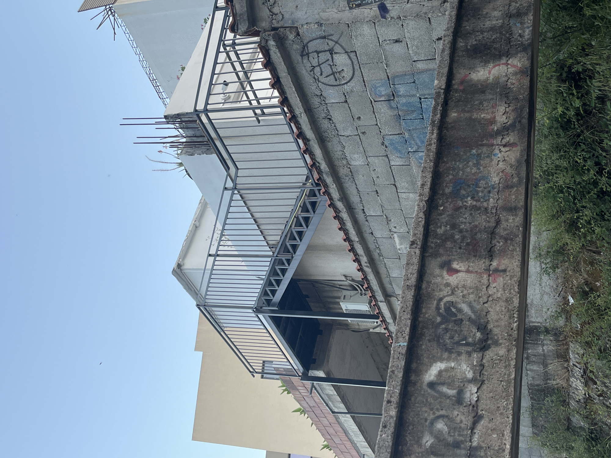 The image depicts an outdoor scene in Budva, Montenegro, characterized by a clear blue sky and a view of urban architecture. In the foreground, a concrete structure, possibly a bridge or overpass, is covered in graffiti. The graffiti consists of various symbols and markings in black, red, and blue. Behind this, a rough concrete block wall is also adorned with some graffiti, including a circular symbol with lines inside. A roof with red tiles is visible atop this wall.
Further back, an external metal staircase with a black railing ascends towards a building. This staircase appears to be part of a modern addition or renovation. Adjacent to the staircase, an outdoor air conditioning unit is mounted on a wall. In the background, several buildings with different architectural styles are visible, one being a light-colored facade and another a stark white structure. The overall environment suggests a partially developed or older urban area. A small, dark object is visible in the sky, but its nature is unclear. Lush green vegetation grows along the bottom of the scene.