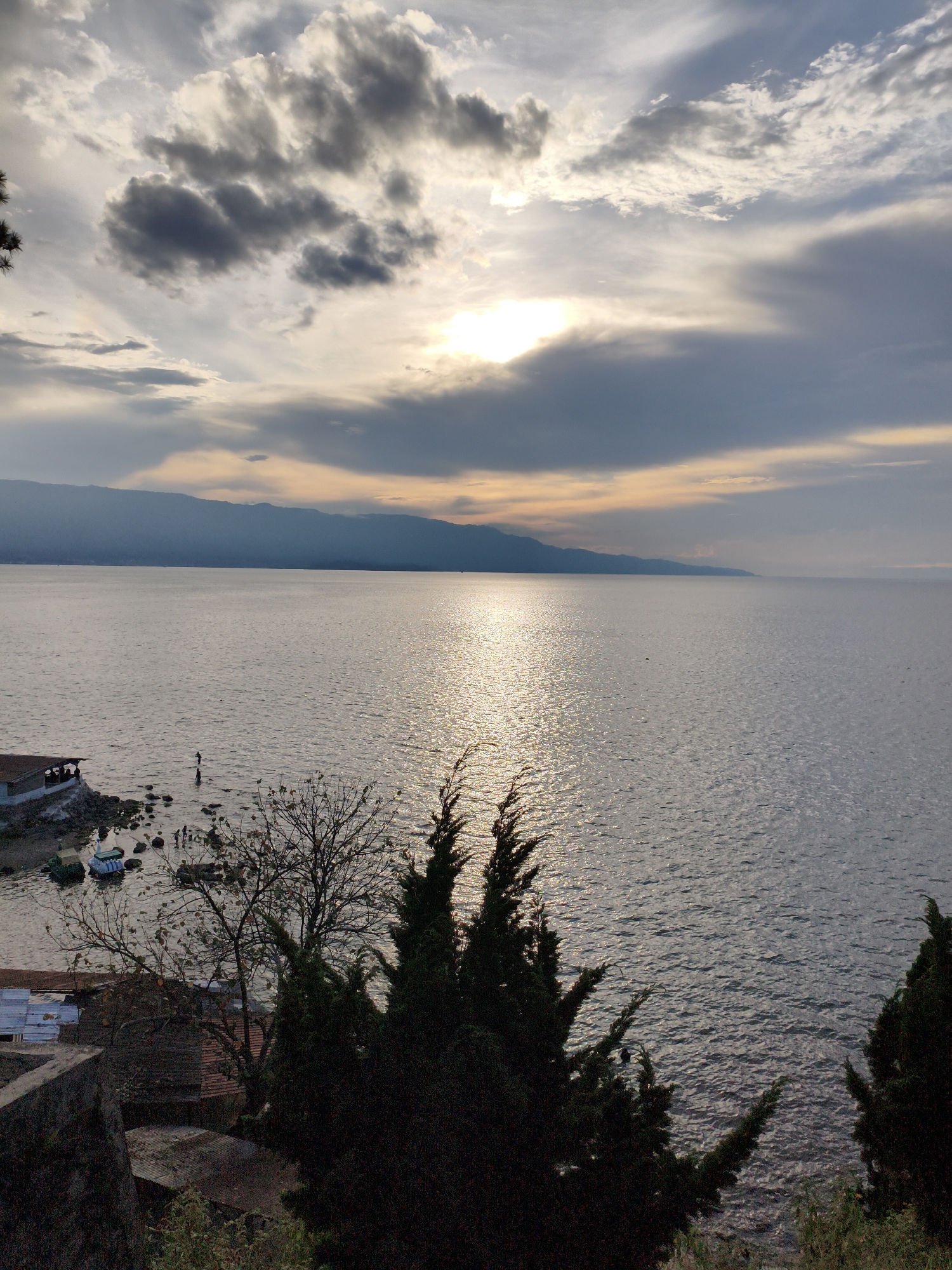 This image captures a serene sunset over a calm body of water, likely a lake or sea, with a distant mountainous shoreline. The sky is a canvas of dramatic clouds, some dark and heavy, others illuminated by the setting sun, which casts a shimmering, golden reflection across the water's surface.
In the foreground, dark silhouettes of evergreen trees frame the scene, their branches stark against the lighter sky. Below them, on the shore, are a few small buildings with corrugated metal roofs, and several boats moored near the rocky edge. A small group of people are visible near the water's edge, some wading, others standing. The overall impression is one of tranquil, natural beauty tinged with human presence.
The location is stated to be in the city of Tiga Raja, Indonesia. This likely refers to Tiga Raja village on Samosir Island, Lake Toba. Lake Toba is a large volcanic lake in North Sumatra, Indonesia, known for its stunning natural scenery, including mountains and clear waters, making it a popular tourist destination. The presence of a large body of water, mountains in the distance, and lakeside settlements are consistent with this location. The architecture of the buildings, with their distinct metal roofs, is also typical of rural Indonesian villages.