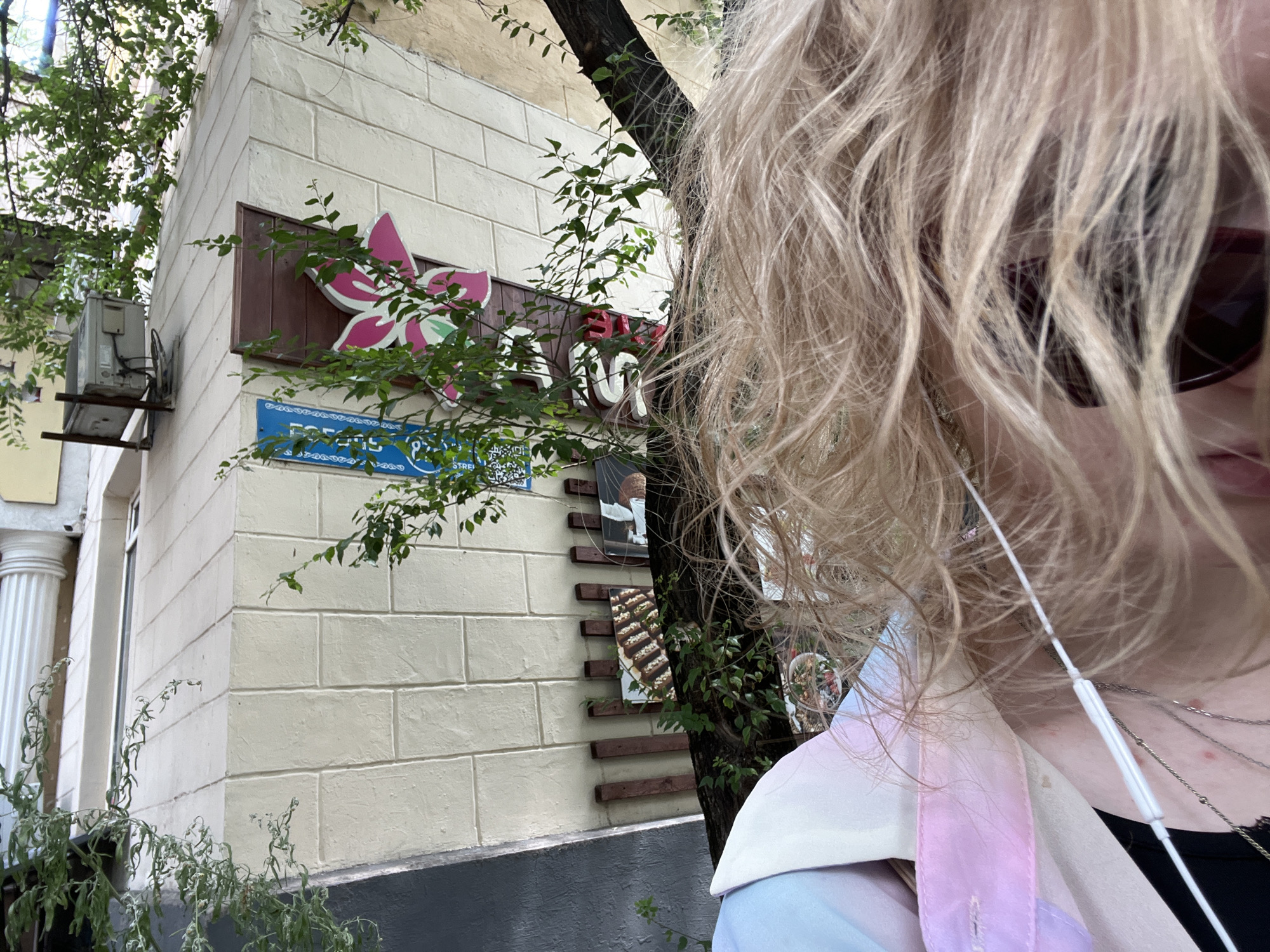 A close-up, selfie-style shot shows a person with blonde, wavy hair in the foreground, obscuring much of their face. They are wearing sunglasses and visible from the chest up. The background features a cream-colored brick building with a tree growing in front of it. A sign with a pink floral design and Cyrillic text is mounted on the building. Below this, a blue sign with more Cyrillic text is visible. Several framed pictures of food are mounted on wooden planks attached to the tree. The overall setting appears to be an outdoor urban environment, likely during the daytime, with dappled sunlight and greenery suggesting a pleasant, possibly warm, day. The presence of an air conditioning unit mounted on the side of the building and a column on the left side of the image hint at a commercial or residential building. The person's attire and the close framing suggest a casual and personal moment, possibly while exploring or commuting. The image conveys a sense of everyday life in an urban setting.