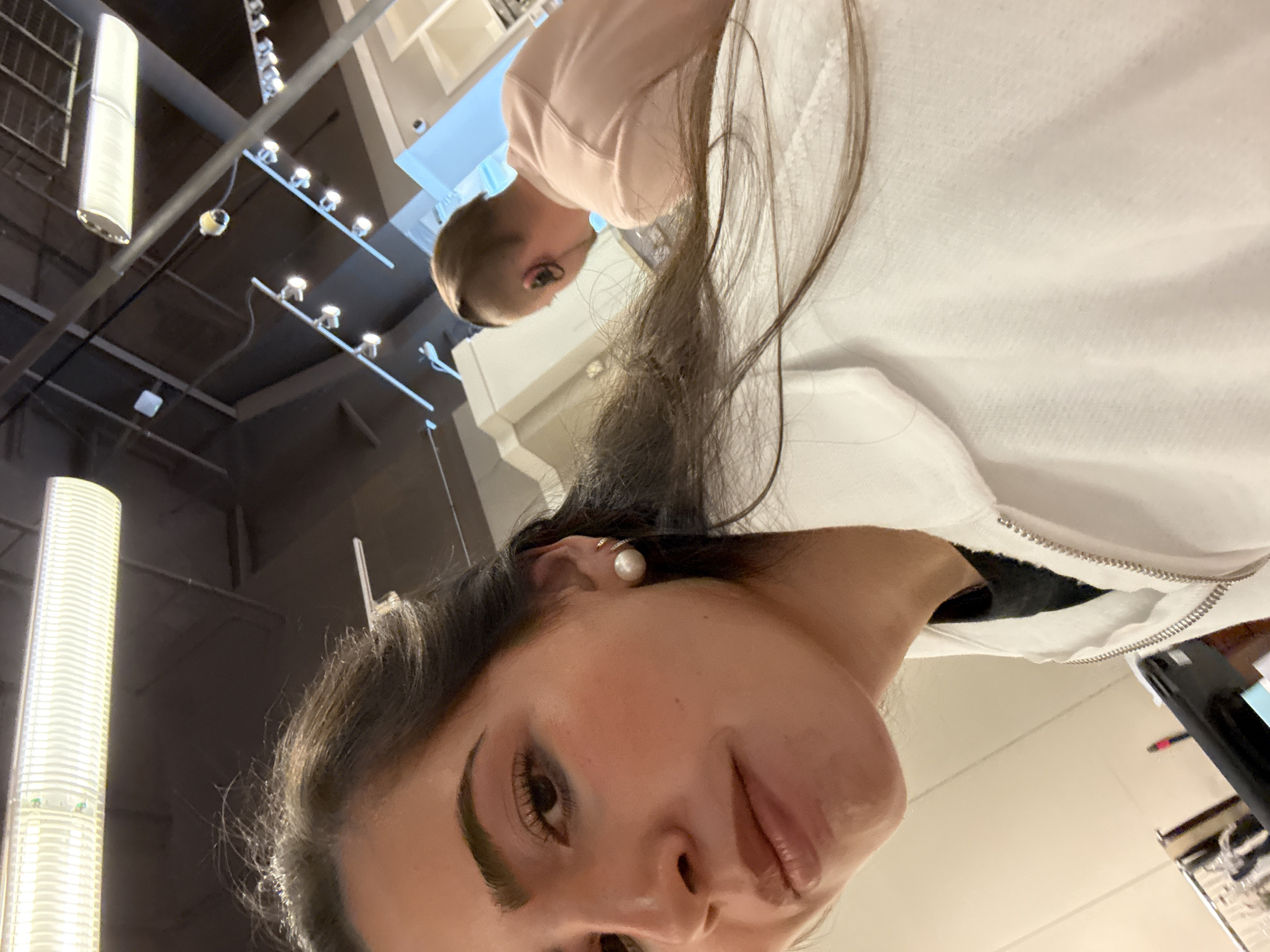 A selfie taken indoors with a woman wearing a white hooded sweatshirt. There is another person in the background wearing a pink shirt. The location appears to be a retail store in San Marcos, Texas, given the ceiling lights and store fixtures.
