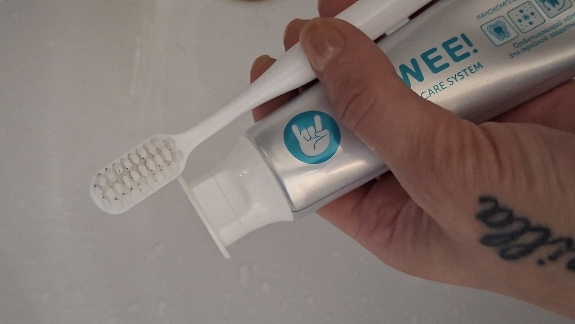 A person's hand holds a white toothbrush and a silver tube of toothpaste. The toothbrush bristles have some dark residue on them. The toothpaste tube has a teal circle with a white hand gesture resembling a rock 'n' roll sign. Text on the tube reads "WEE! CARE SYSTEM". Additional text and icons related to dental care are visible. A tattoo is present on the person's hand. The background appears to be a light-colored, possibly tiled surface, suggesting a bathroom environment. The location is not identifiable from the image.