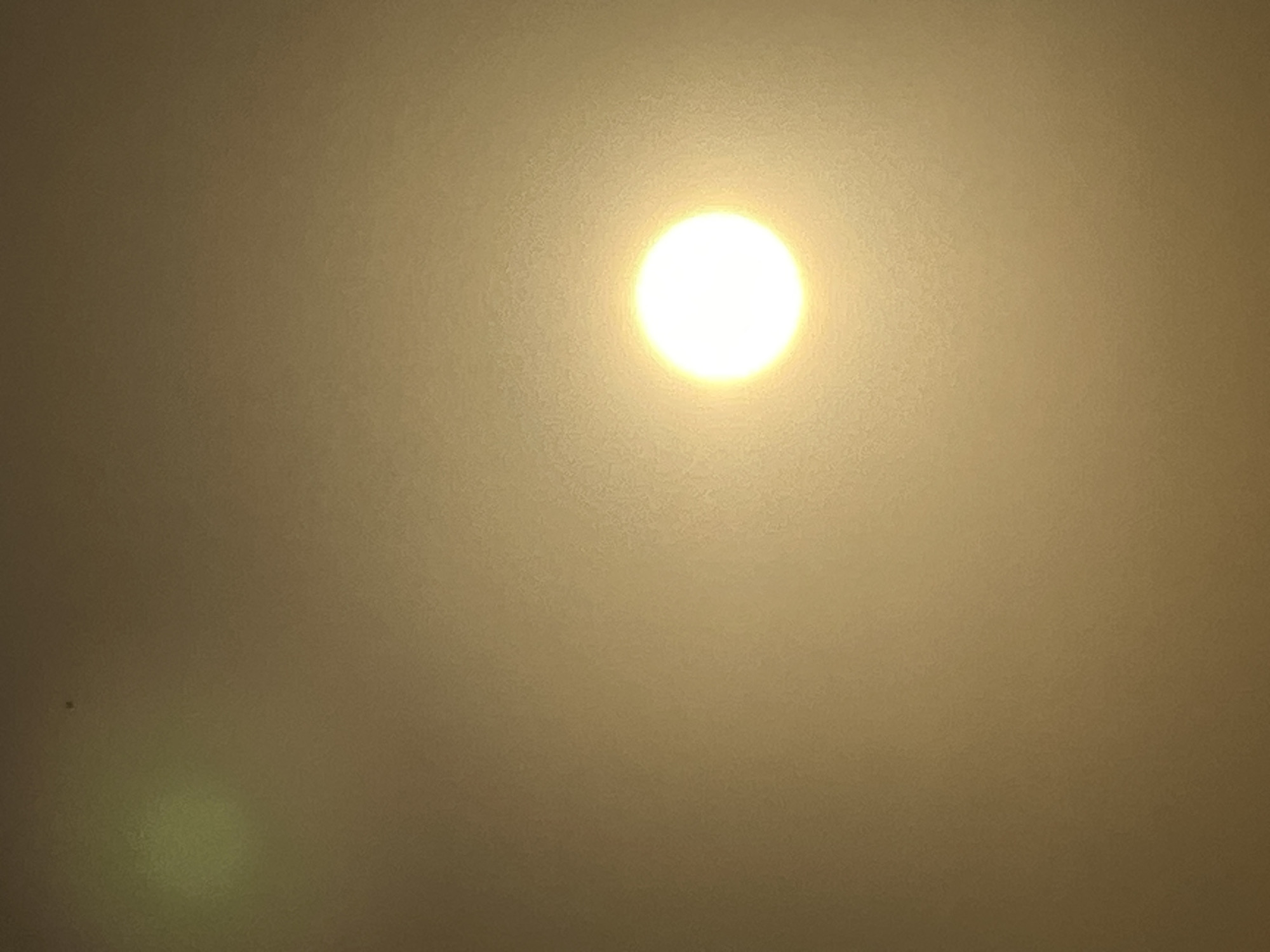 A bright, circular light source dominates the right half of the frame, suggesting a sun or a very strong artificial light. The overall scene is illuminated by a warm, golden glow, creating a hazy or slightly diffused atmosphere. In the upper left quadrant, a fainter, yellowish-green circular area is visible. A tiny, dark speck is discernible in the upper central portion of the image. The background appears to be a textured surface, possibly a wall or ceiling, with a grainy quality. No people or specific urban features are identifiable in this abstract representation of light and atmosphere, so it is not possible to determine if it is located in Kuta, Indonesia.