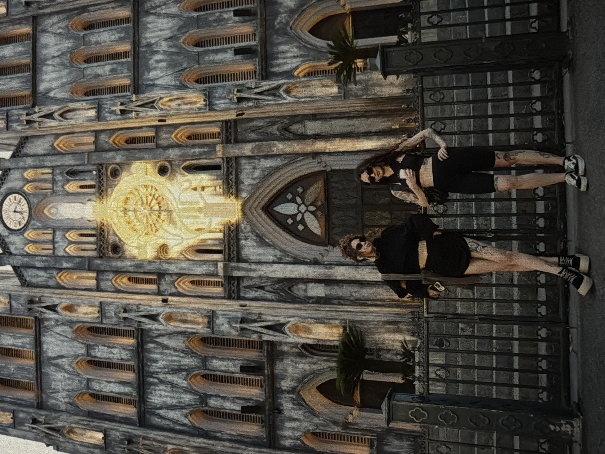 Two women stand on an ornate, dark, metal balcony in front of a large, detailed stone building. The building appears to be a cathedral or similar religious structure, with a clock visible on its facade. The scene is in Da Nang, Vietnam. The woman on top has long dark hair, sunglasses, and wears a black sports bra and bike shorts. She has tattoos on her arms and legs. The woman below has short curly blonde hair, sunglasses, and wears a black long-sleeve shirt and shorts. She also has tattoos on her legs.