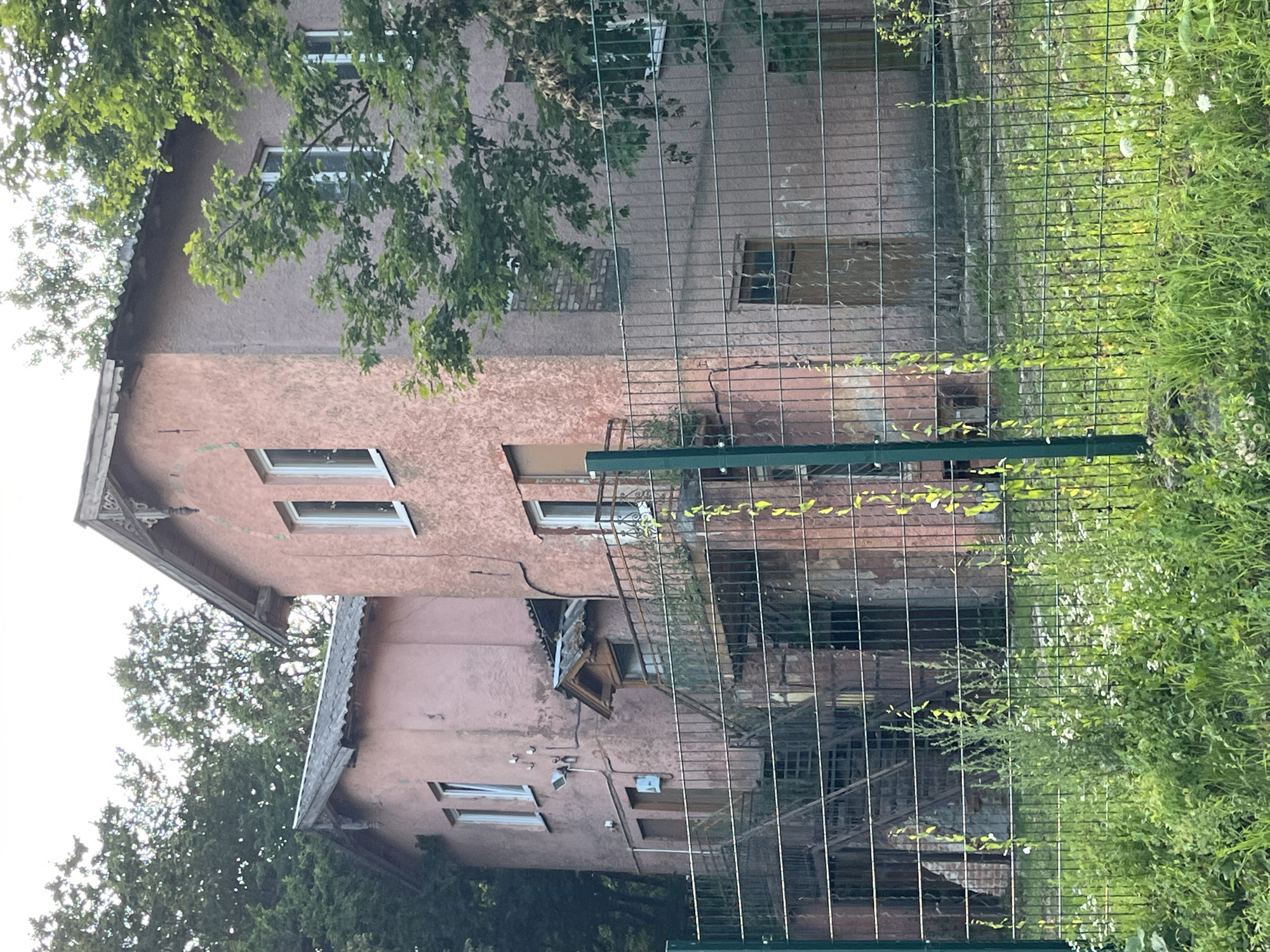 The image shows a two-story pink building with a corrugated metal roof, surrounded by trees. A metal fence runs in front of the building, with green foliage growing in front of the fence. The building appears to be old and possibly abandoned, with some signs of disrepair. The windows are mostly intact, but some appear to be boarded up. There is a balcony on the second floor of the building, accessible by an outdoor staircase. The overall scene is one of neglect and decay, set against a backdrop of lush green vegetation. The location is identified as Svetlogorsk, Russia.