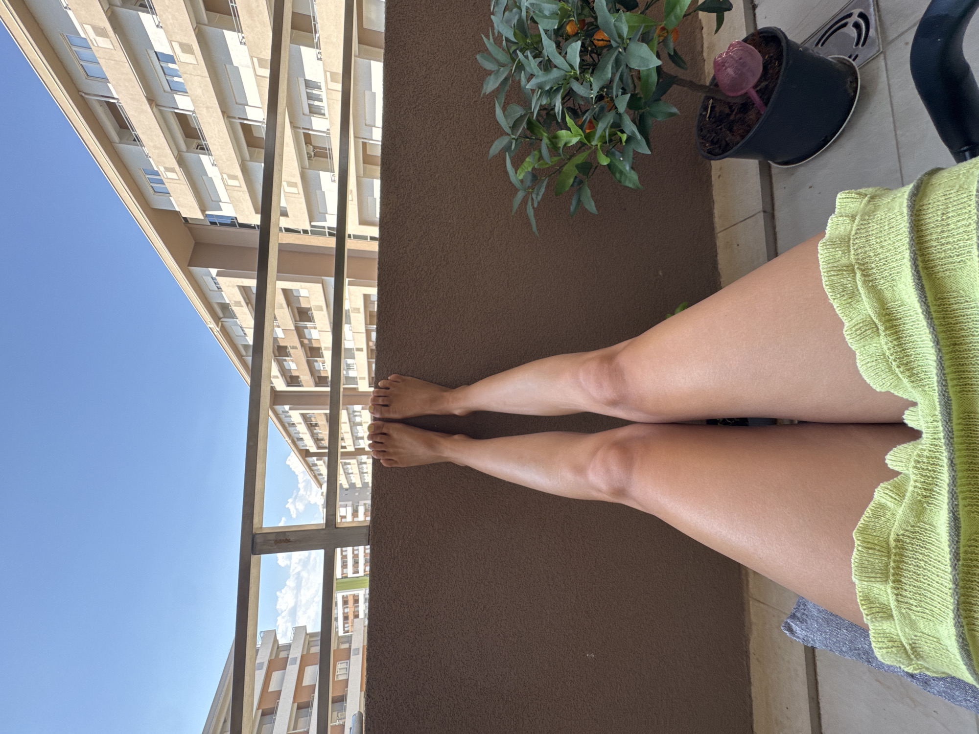 A pair of bare human legs, positioned vertically in the frame, rests against a brown wall. The legs are tanned, and the individual is wearing a bright yellow garment with a ruffled hem. To the right, a potted plant with small orange fruits sits on the tiled balcony floor. In the background, apartment buildings with light-colored facades and balconies are visible under a clear blue sky. A metal railing separates the balcony from the view of the buildings. The scene appears to be a casual moment on a sunny day.