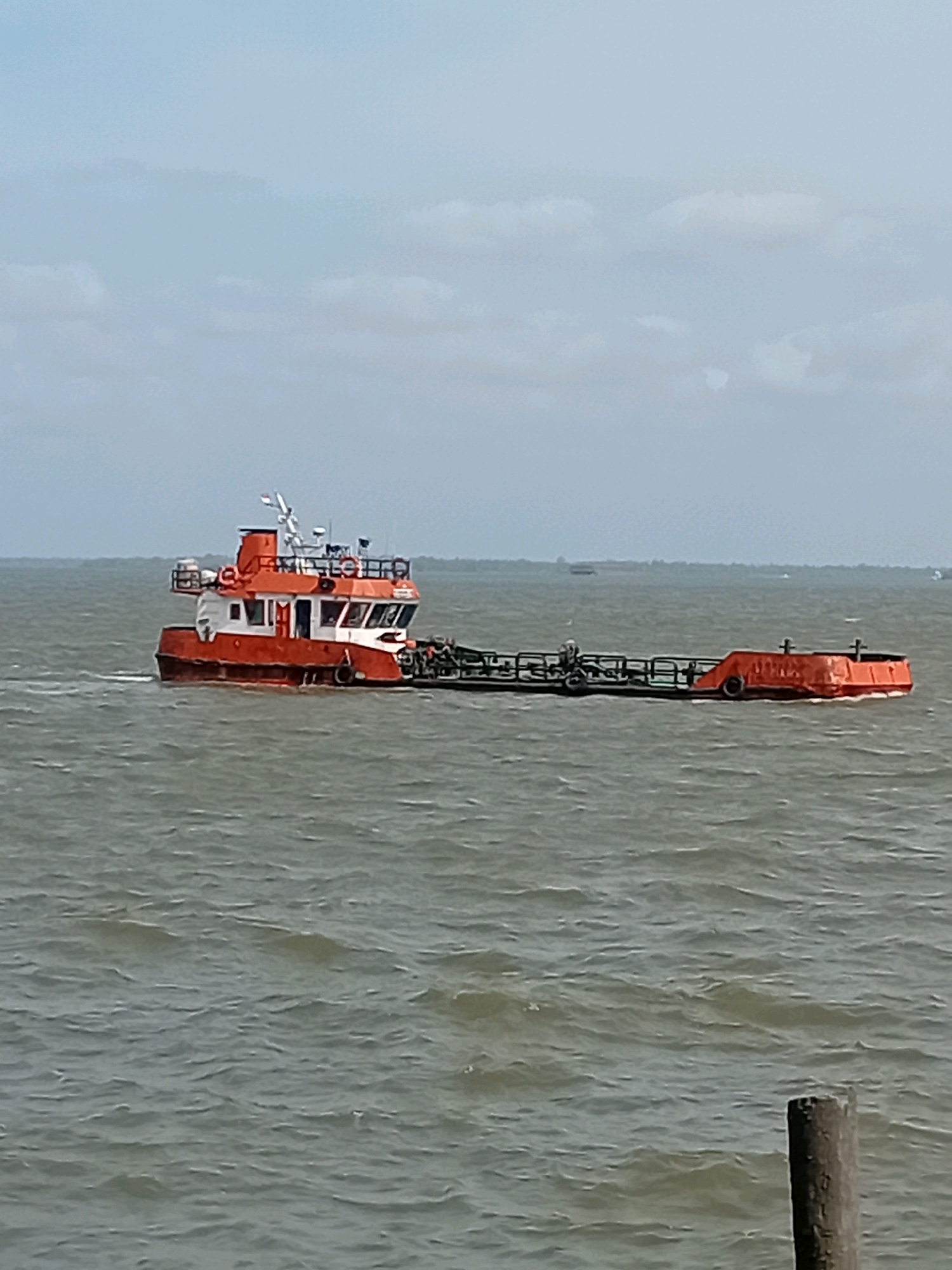 A small orange and white tugboat is pushing a long, flat barge on choppy, grey water under an overcast sky. The tugboat has a white cabin with dark windows and an orange roof. The barge is black with orange ends and appears to be carrying some equipment. In the distance, a faint treeline is visible on the horizon. The scene is located in the waters off Sungai Meriam, Indonesia. There are no people visible in the image.