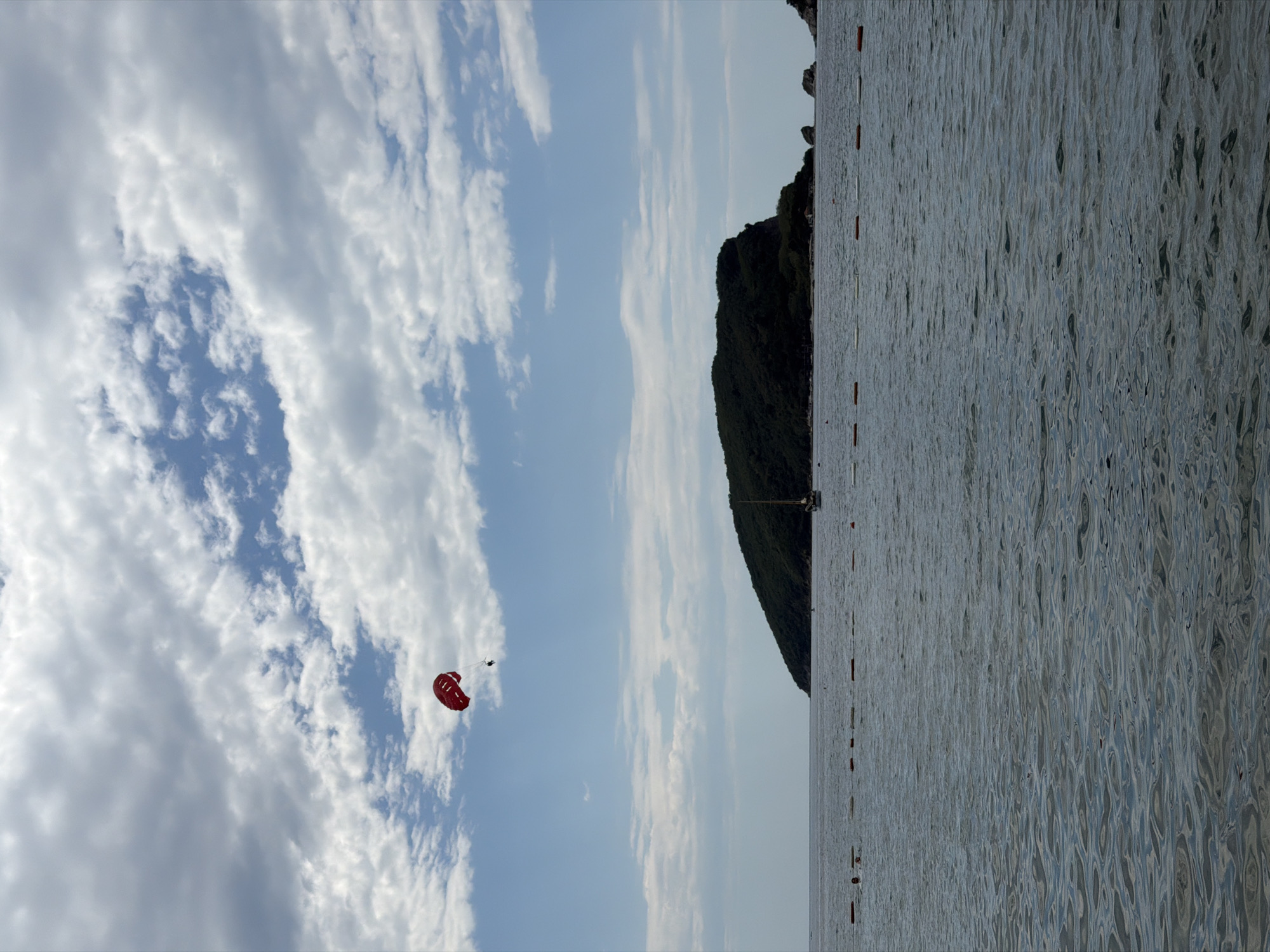 The image captures a sunny day at a beach in Budva, Montenegro. The horizon is somewhat obscured by a lush, green hill rising from the sea. To the left, the sky is filled with puffy white clouds, giving way to a blue sky in the center of the frame. A red parachute with a figure attached is visible against the cloudy sky. The sea is calm with subtle ripples and reflections, taking up most of the right portion of the image. Several small, orange buoys line the water's edge near the hill.