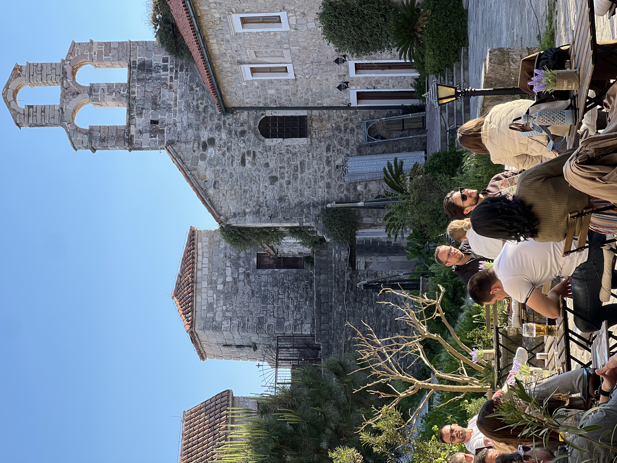 The image depicts an outdoor café scene adjacent to historic stone buildings in Budva, Montenegro. A group of people are seated at wooden tables, some engaged in conversation. A man in a white shirt is sitting at a table with a glass of beer. The architecture features a stone bell tower with arched openings and buildings constructed with light-colored stone. The café is bordered by greenery, including trees and potted plants. The sky is clear and blue. A lamp post is visible near the tables.