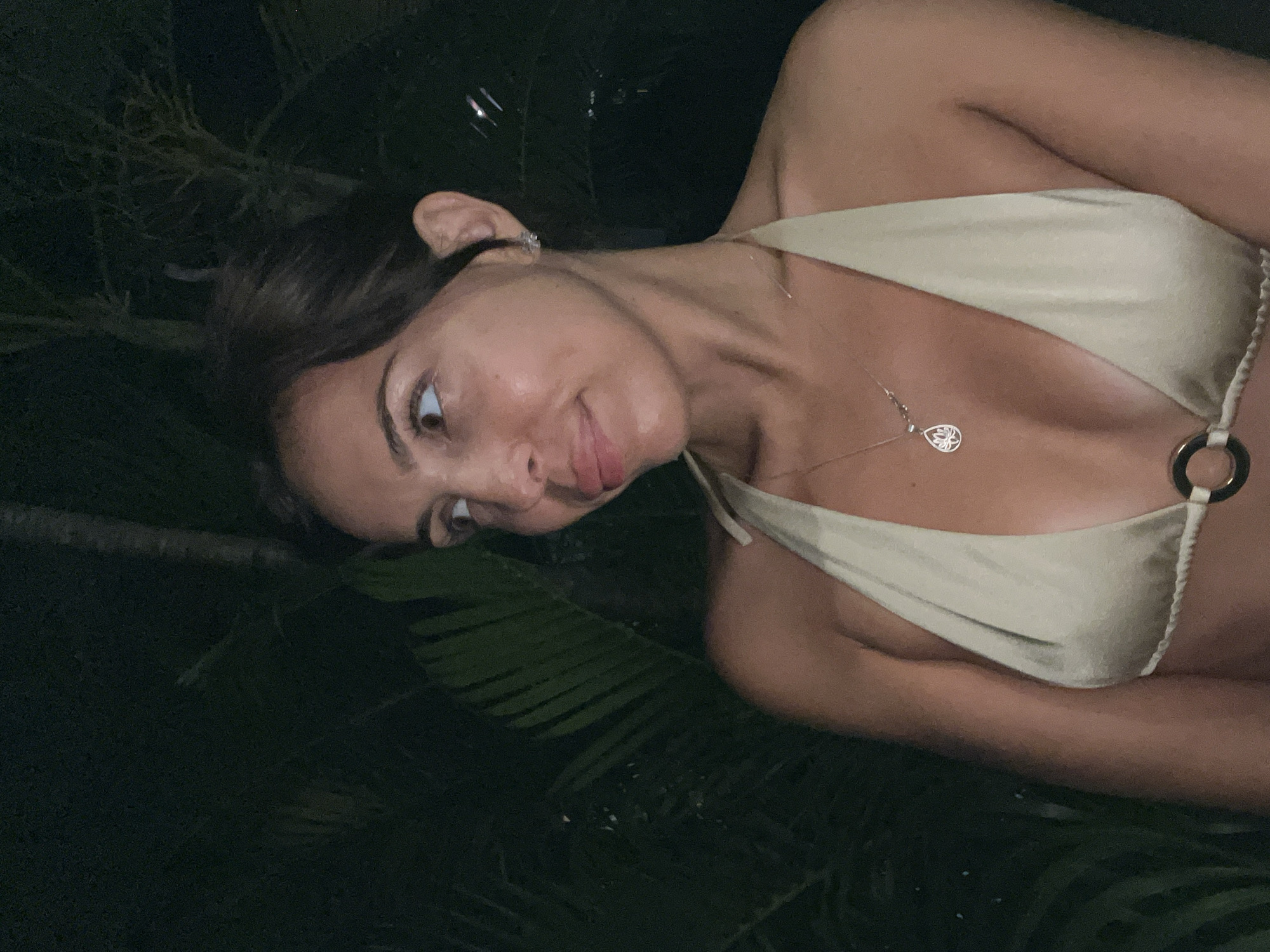 A young woman is shown in a dimly lit outdoor scene, likely at night. She is positioned diagonally in the frame and wearing a light-colored halter bikini top and a delicate necklace with a pendant. Her dark hair is pulled back, and she is looking toward the viewer with a slight smile. The background appears to be foliage, including what seem to be palm leaves. The location is inferred as Kuta Selatan, Indonesia. The image appears to be a casual photograph.