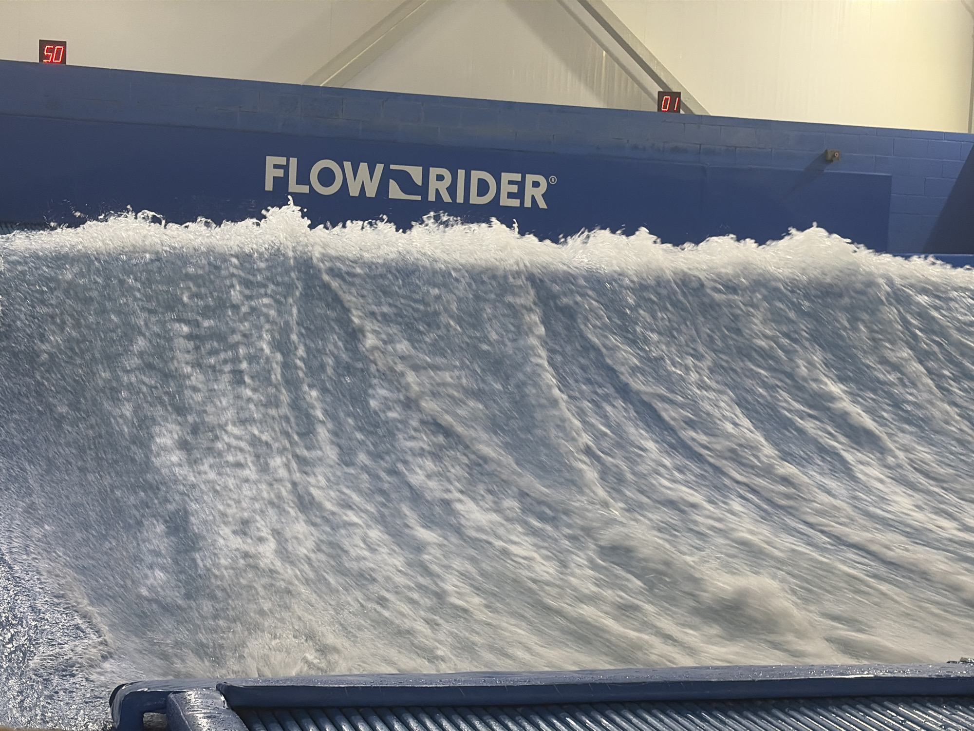 An indoor FlowRider surf simulator in Lake Delton, Wisconsin, displays a large, powerful artificial wave. The wave is in motion, appearing as a sheet of white water cascading down a textured surface. Above the wave, a blue barrier with the white "FLOWRIDER" logo is visible. Digital displays show "50" on the left and "01" on the right, likely indicating time or score. The environment appears to be an indoor facility with a white ceiling. A dark blue, ribbed structure is in the foreground, at the base of the wave.