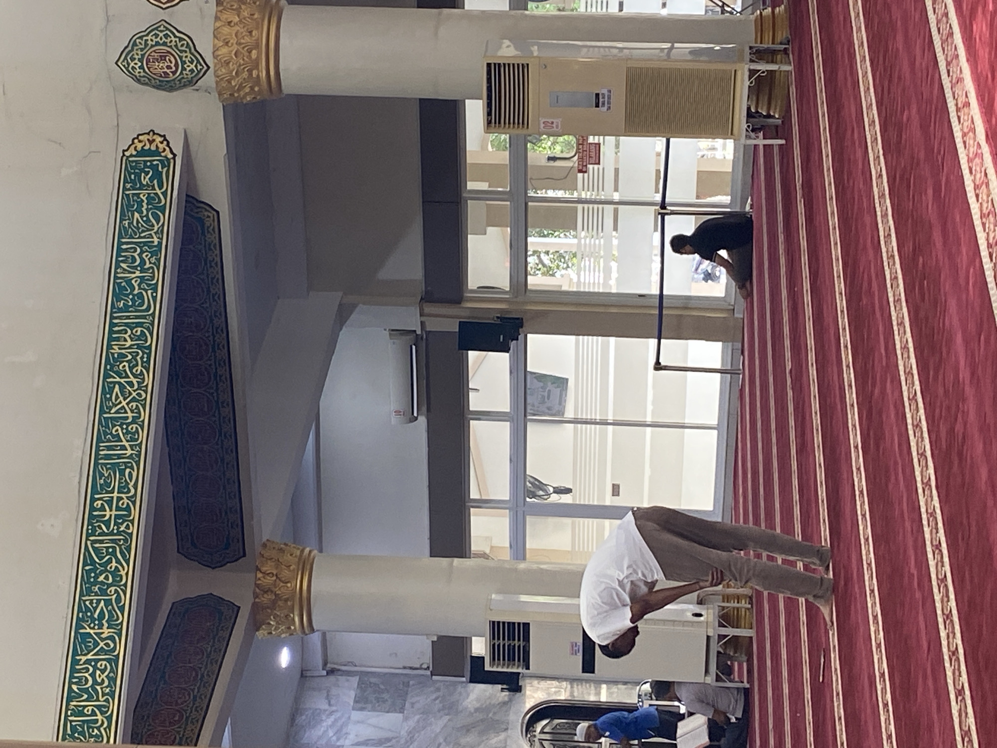 In Lhokseumawe, Indonesia, the image captures a mosque interior with worshippers. A man in a white shirt and brown pants is bowing during prayer, facing a red carpet with striped patterns. Another individual kneels further away, near a window. The mosque features white columns with ornate gold details, and walls adorned with green and gold Islamic calligraphy. Large windows provide natural light, and air conditioning units are visible. The floor is tiled with a marble-like pattern.