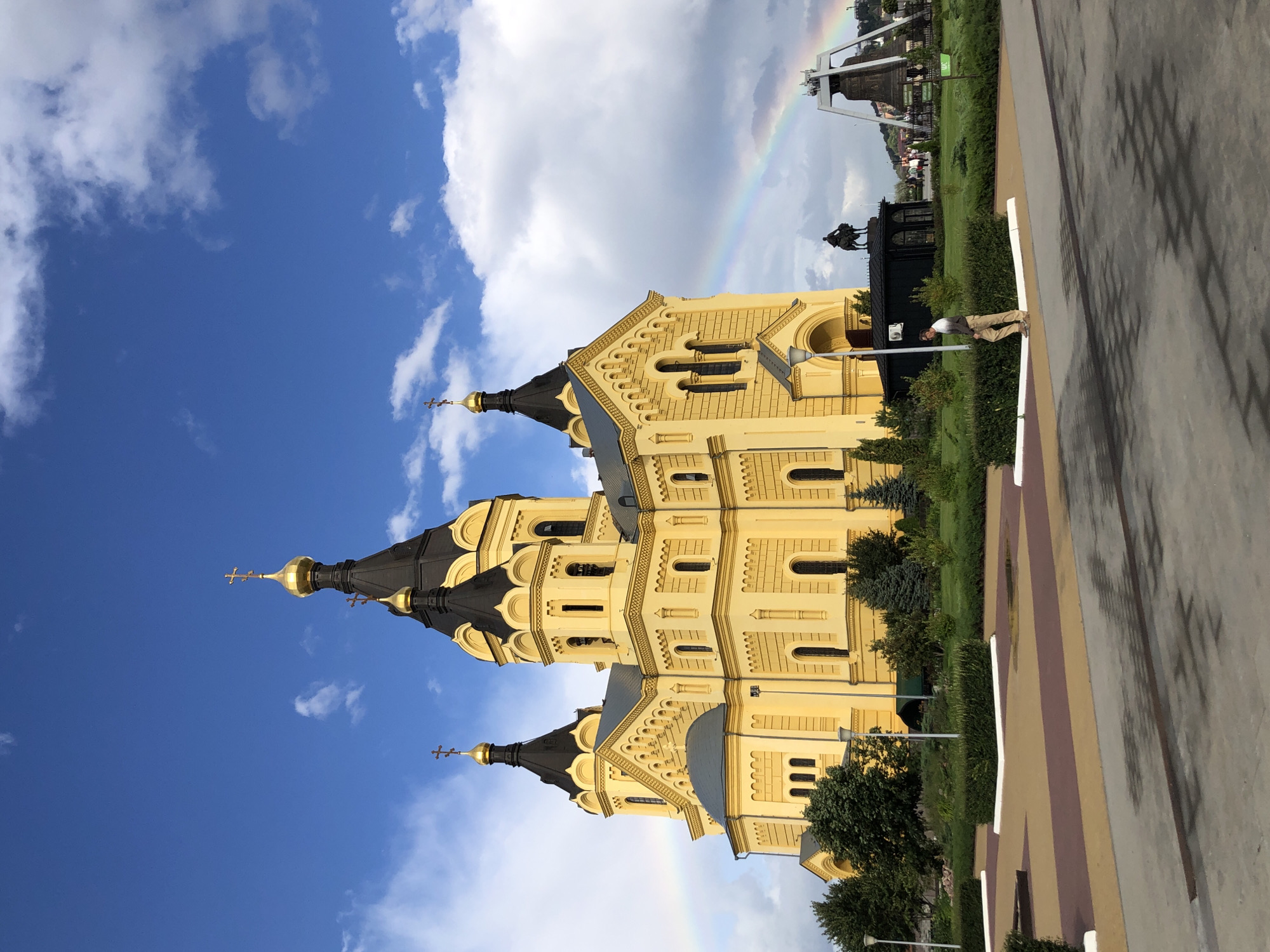 The image depicts the Alexander Nevsky Cathedral in Nizhny Novgorod, Russia, on a partly cloudy day with a rainbow visible in the sky. The grand yellow cathedral, with its black domed roofs and golden crosses, dominates the left side of the frame. In the foreground, a paved area with alternating colored sections leads towards a neatly landscaped garden with hedges and trees. A single person, wearing light-colored pants and a striped shirt, walks along the edge of the garden. To the right of the cathedral, a large bell is displayed within a metallic structure, and further in the background, a statue of a man on horseback can be seen. The overall scene conveys a sense of calm and grandeur, highlighting the architectural beauty of the cathedral and the surrounding public space.