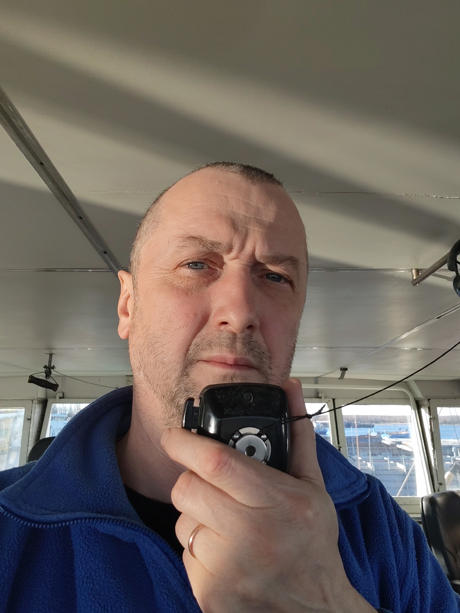 A close-up, eye-level portrait features a man, likely middle-aged, with a bald or closely-shaven head, blue eyes, and light facial stubble. He is wearing a dark blue fleece jacket over a black shirt, and a plain gold ring is visible on the ring finger of his left hand. The man is holding a black two-way radio or walkie-talkie to his mouth with his right hand, indicating he is speaking or listening. The radio has a silver circular speaker or control on its face and a thin black cord.
He looks directly at the camera with a serious or thoughtful expression, his brow subtly furrowed. The setting is the interior of what appears to be a boat or ship's cabin or bridge. The light-colored ceiling, possibly white or light grey, features visible metal beams or framing and a horizontal line of small fasteners or rivets. Bright daylight streams in from large windows in the background, illuminating the scene and casting some shadows. Through these windows, calm water is visible, along with several boats or dockside structures in the distance under a clear, bright sky, suggesting a sunny day in a harbor or port. The image depicts a man likely at work in a maritime environment. No text is visible in the frame.