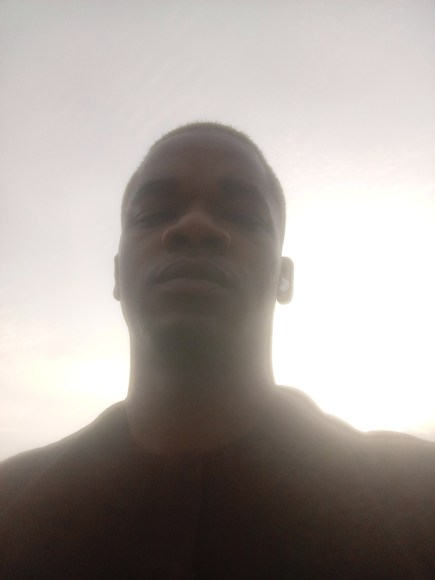 A close-up, slightly blurry, selfie-style photograph shows the upper body and head of a person, presumably male, against a bright, overcast sky. The subject is facing the camera directly, with their face angled slightly upwards. The lighting is very bright and appears to be coming from behind the subject, causing overexposure in the upper portion of the image and obscuring details in the background. The subject's skin tone is dark, and they have a buzz cut hairstyle. Their facial features are discernible, including their nose, mouth, and ears. The subject is wearing a dark-colored shirt or t-shirt. The overall impression is that the photograph was taken outdoors, possibly during daylight hours, with the sun positioned behind the subject or the sky being very bright. The context provided places this image in Gwarinpa, Nigeria.