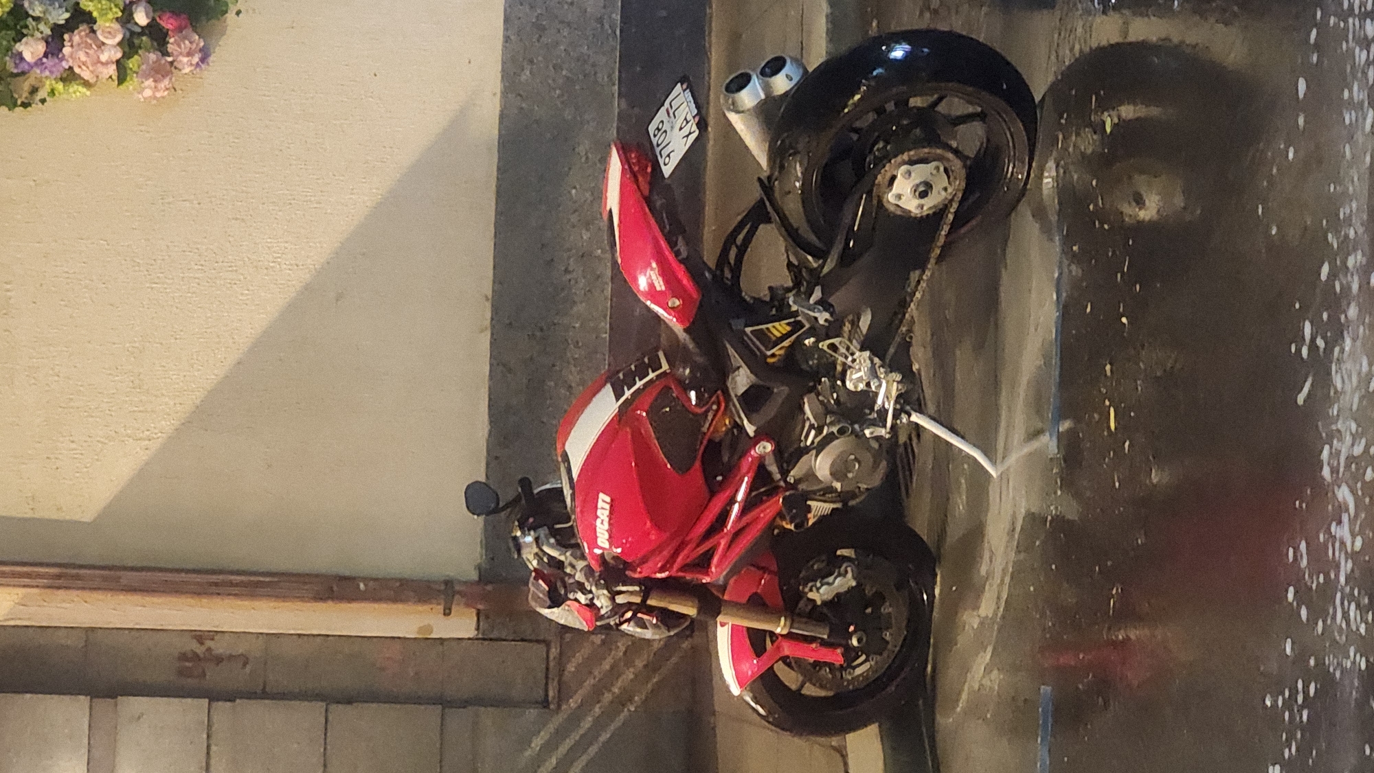 A red Ducati motorcycle is parked on a street. The motorcycle has a license plate that reads "9708 XA 77." The location is Moscow, Russia. The street surface appears wet. There are flowers in the upper left corner of the image. The scene appears to be at night.