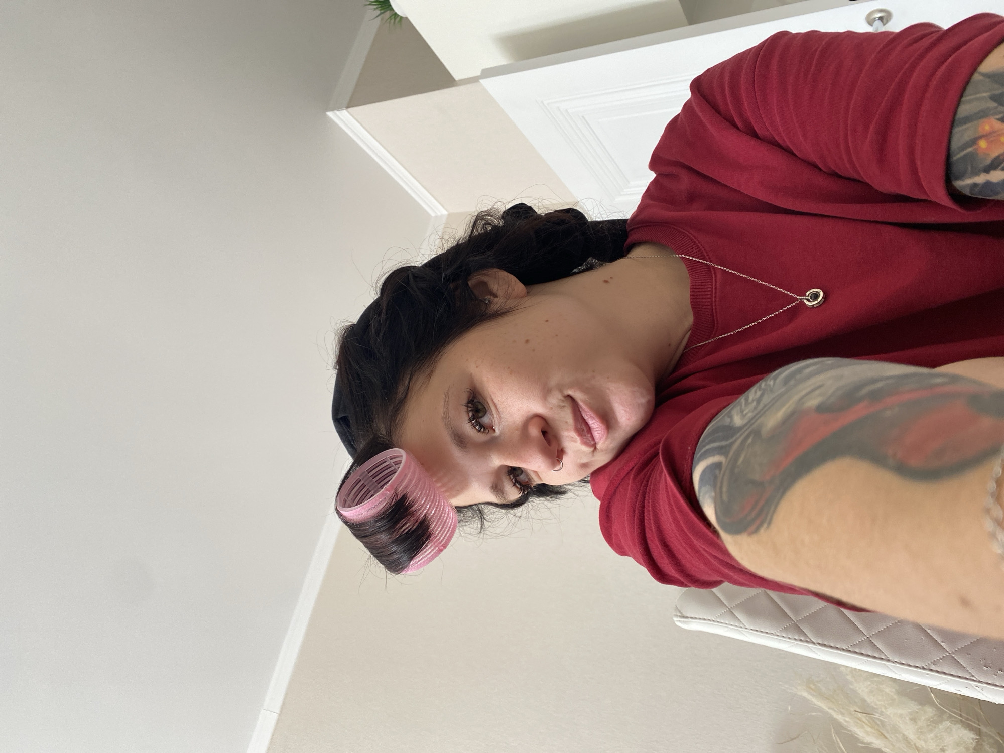 A young woman with dark hair styled with pink hair rollers is pictured in an indoor setting. She is wearing a red sweatshirt and a silver necklace. Her right arm, adorned with a tattoo, rests on her shoulder. The background features a white door and a wall. The image was taken in Yanino-1, Russia.