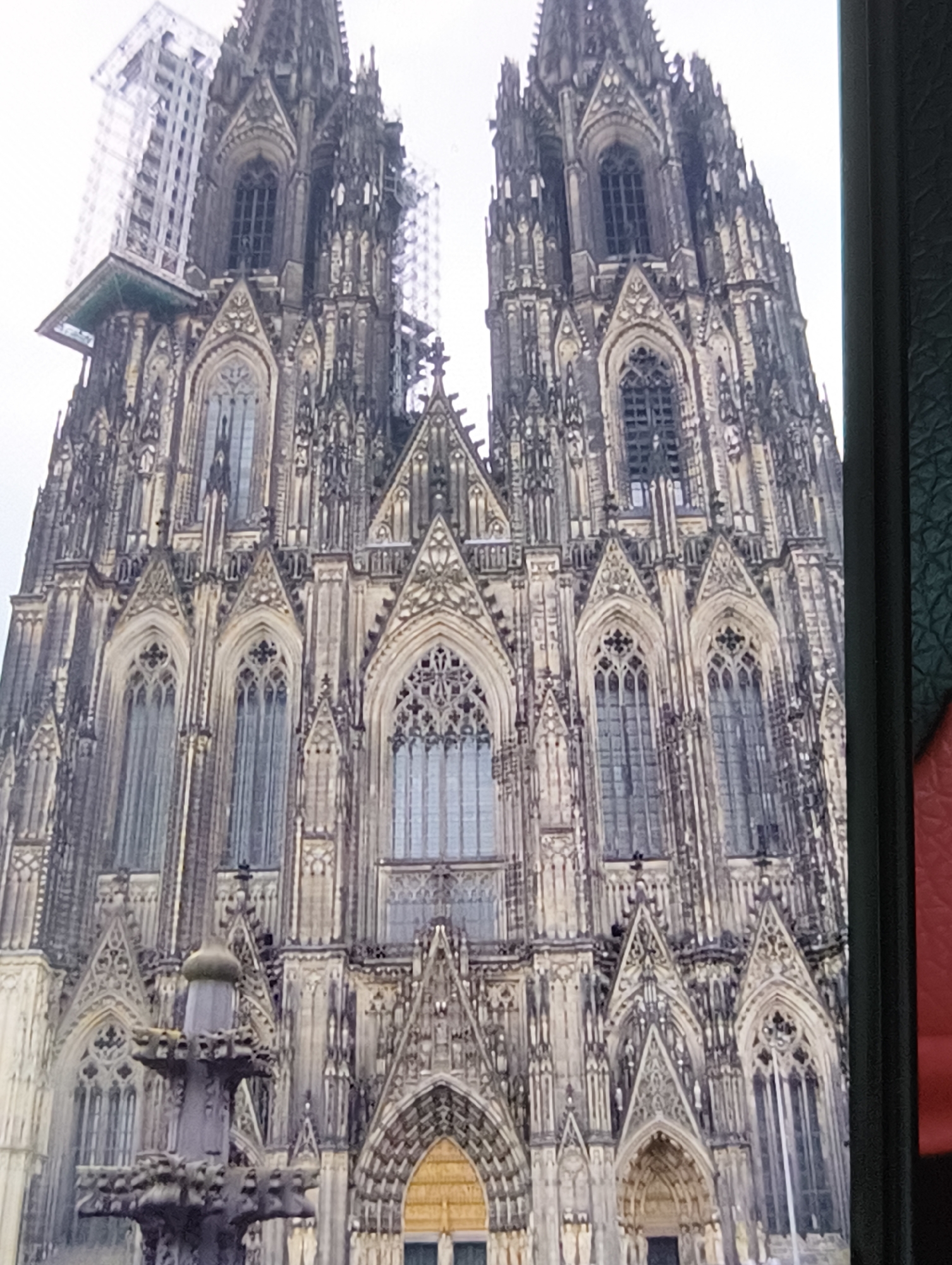 The image displays a close-up, vertical view of a large Gothic cathedral. The cathedral's facade is intricately detailed with numerous spires, arched windows with tracery, and decorative stonework. Two prominent towers with pointed spires dominate the upper part of the image. In the foreground, a portion of a dark, textured object, likely the edge of a device or frame, is visible on the right side. The sky appears overcast and bright. Construction or scaffolding is partially visible on the left side of the upper facade. A dark, ornate fountain-like structure is situated in the lower left quadrant. The overall impression is one of grand architecture and intricate craftsmanship.