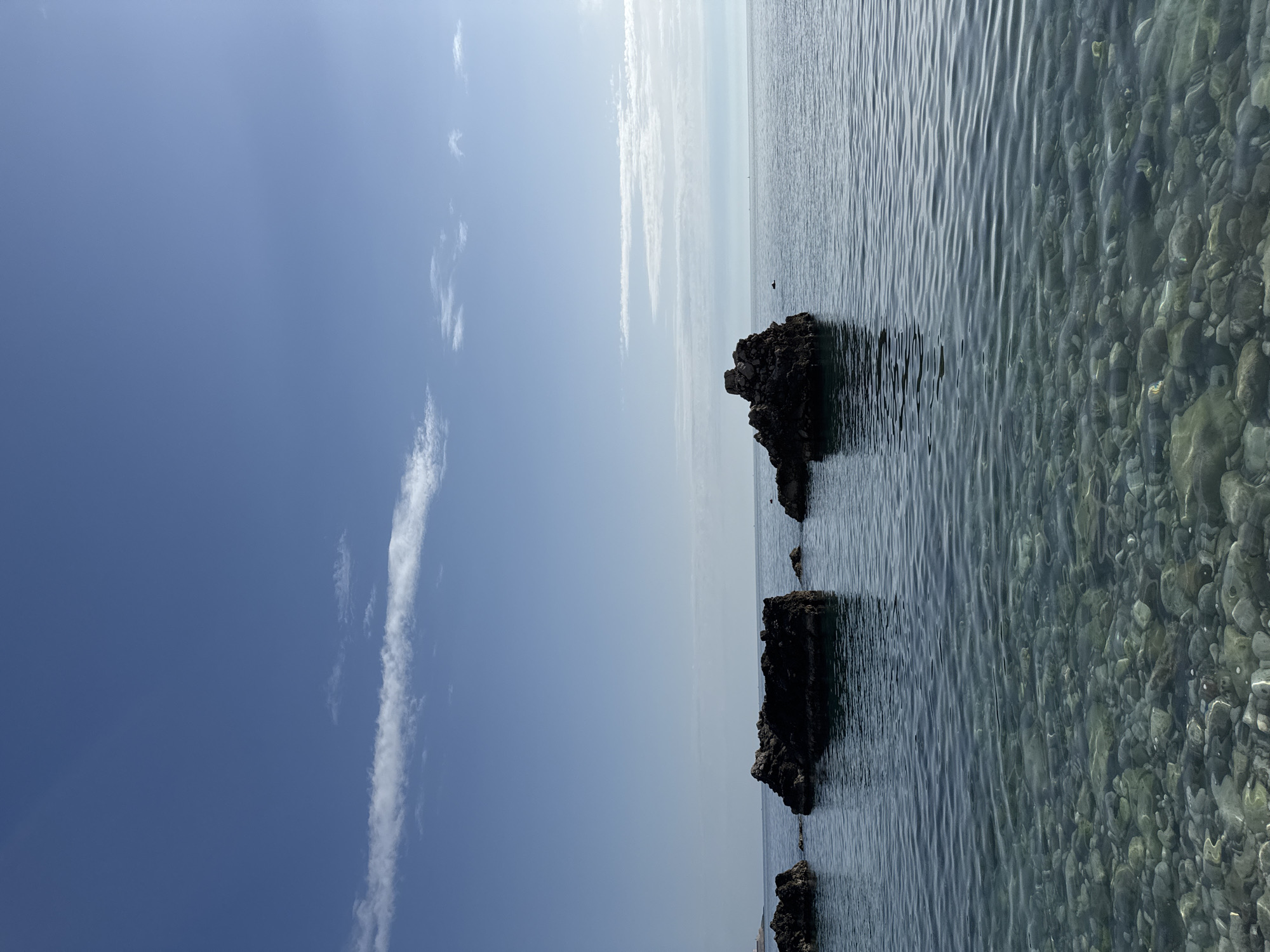 The image shows a view of the sea from a beach in Bar, Montenegro. The sky is mostly clear with a few elongated clouds. The water is calm and clear, revealing pebbles on the seabed near the shore. There are several dark, rocky outcrops extending into the sea. The location is the city of Bar, Montenegro.