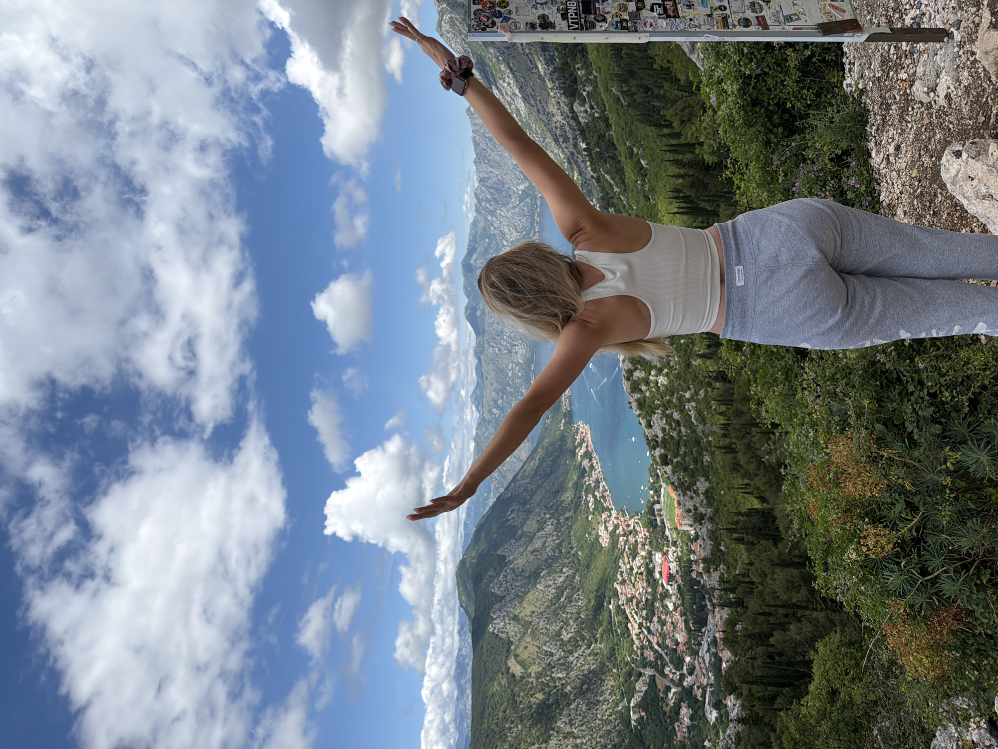 A blonde woman, seen from behind, stands at a mountain viewpoint with her arms outstretched horizontally, seemingly embracing the panoramic landscape. She wears a white ribbed crop top, grey Calvin Klein sweatpants with the brand name visible on the waistband tag and down the left leg, a scrunchie, and an Apple Watch on her left wrist.
The setting is a high vantage point overlooking a dramatic natural scene in Špiljari, Montenegro. Below the woman, steep, lush green mountains covered in trees descend towards a large, calm blue bay or fjord. Along the water's edge, a town or city with numerous buildings is nestled between the mountains and the sea. Distant mountain ranges complete the backdrop under a bright blue sky dotted with scattered, fluffy white clouds.
To the woman's right, a wooden signpost or pillar is visible, heavily adorned with various stickers, some featuring text such as "VTPNB." The ground around her is rocky and uneven, with sparse vegetation. The scene is brightly lit, indicating a sunny daytime. The overall impression is one of freedom, appreciation for nature, and a breathtaking vista.