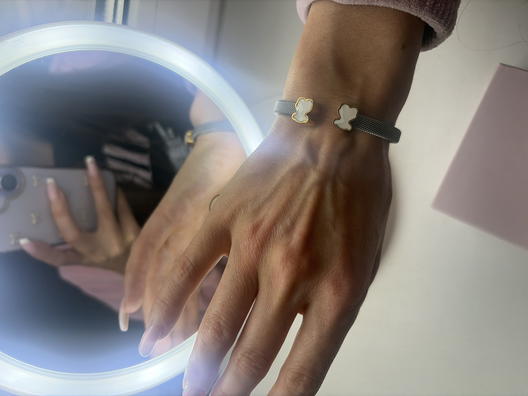 A close-up shot depicts a person's left arm and hand wearing a grey metal mesh bracelet with two bear-shaped charms, one on each end of the bracelet. The skin tone appears light. The person is holding a smartphone, visible in a ring light reflection. The background is mostly blurred, with a white surface and a hint of pinkish-beige. The location is unknown, and there's no information to suggest it's Kudrovo, Russia.