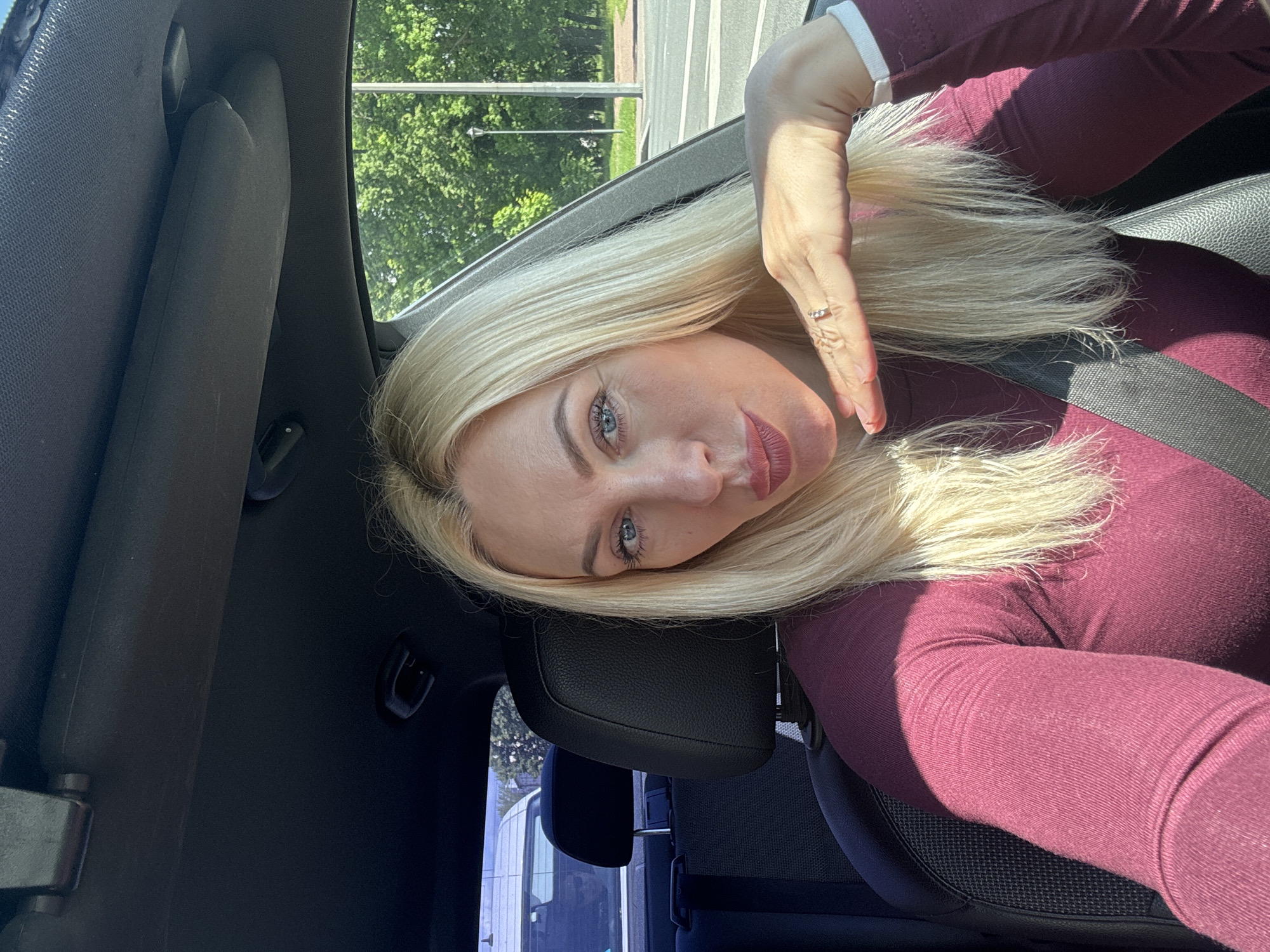 A woman with blonde hair and blue eyes, wearing a burgundy long-sleeved shirt, sits in the driver's seat of a car. She is looking at the camera with her lips pursed and her right hand resting on her chin, adorned with a ring. Sunlight casts shadows on her face and upper body. The interior of the car is visible, including the headrest of the passenger seat and a seatbelt. Through the car window, a blurred background shows green trees, a street, and a lamppost, suggesting an outdoor urban setting.