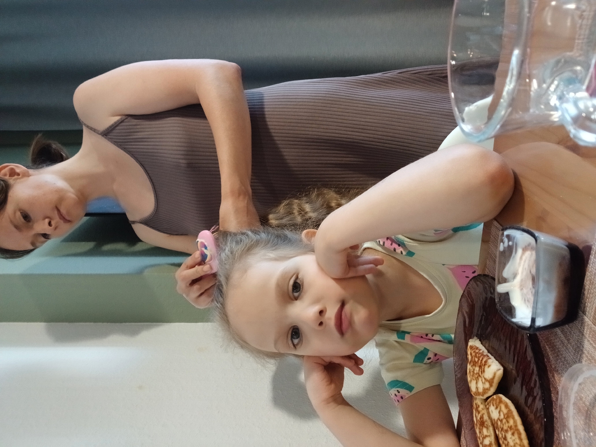 The image shows a woman and a young girl at a table. The woman is wearing a grey ribbed tank top and is leaning over, her arm supporting her head. Her hair is dark and pulled back. The girl, who appears to be around 4-5 years old, is in the foreground, leaning on her hands. She has blonde hair and is wearing a light-colored t-shirt with a watermelon print. She is looking directly at the camera with a curious expression. A pink hair clip is visible in her hair. On the table in front of the girl is a wooden plate with several small pancakes and a small glass dish containing what appears to be whipped cream or yogurt. A clear glass is also visible on the table. The background is a mix of a grey wall and a curtain. The lighting suggests it might be daytime. The overall scene is informal and appears to be a casual meal or snack time. The image is oriented such that the subjects are shown from a sideways perspective. There is no visible text in the image. The location context is Yekaterinburg, Russia.
