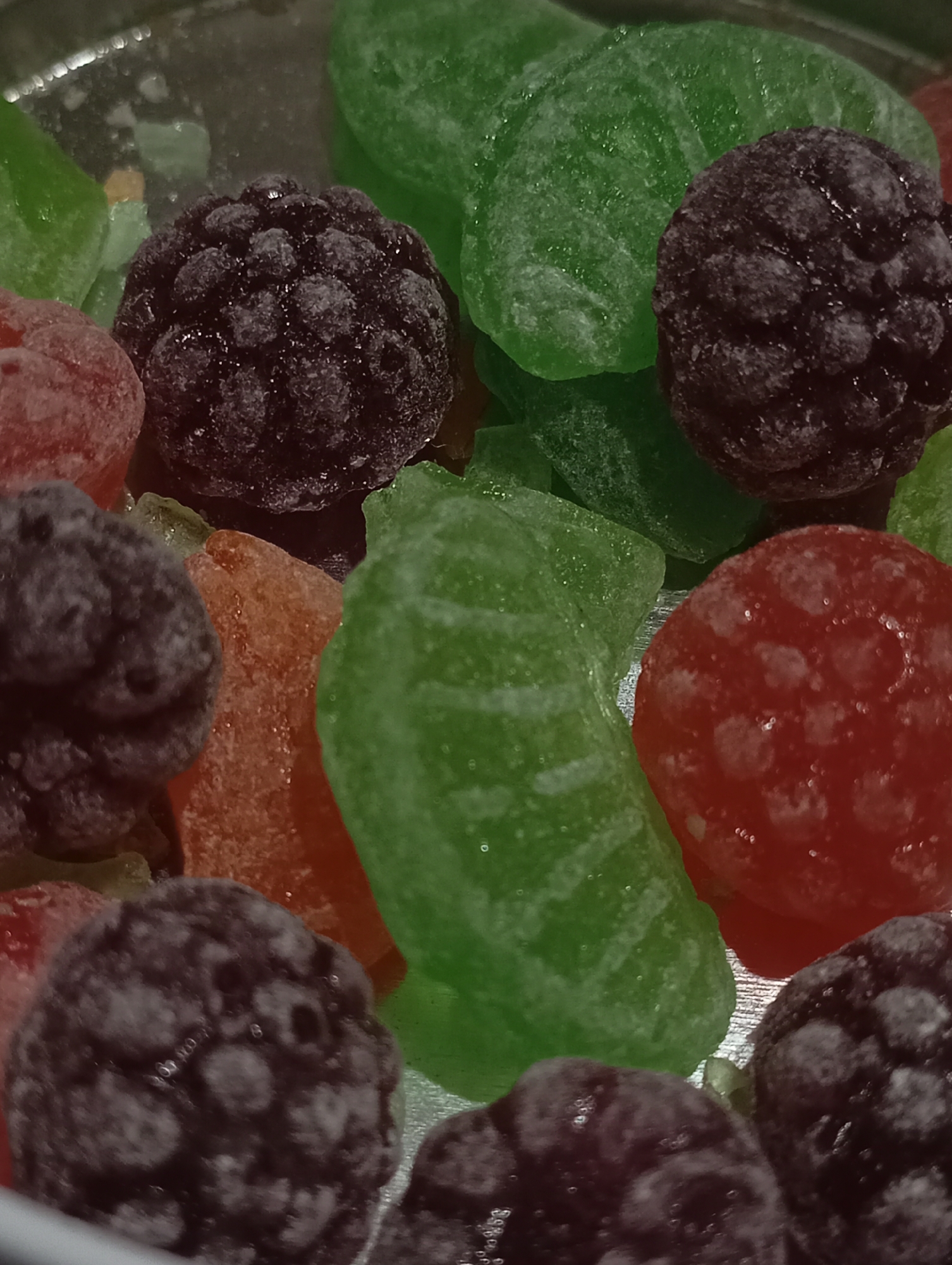 The image shows a close-up of assorted candies. There are several pieces of green, red, and orange hard candy, along with what appear to be sugared blackberries. The candies and blackberries are arranged in a somewhat chaotic manner, obscuring parts of each other. The surface they are on appears to be metallic. No information on location is discernible.