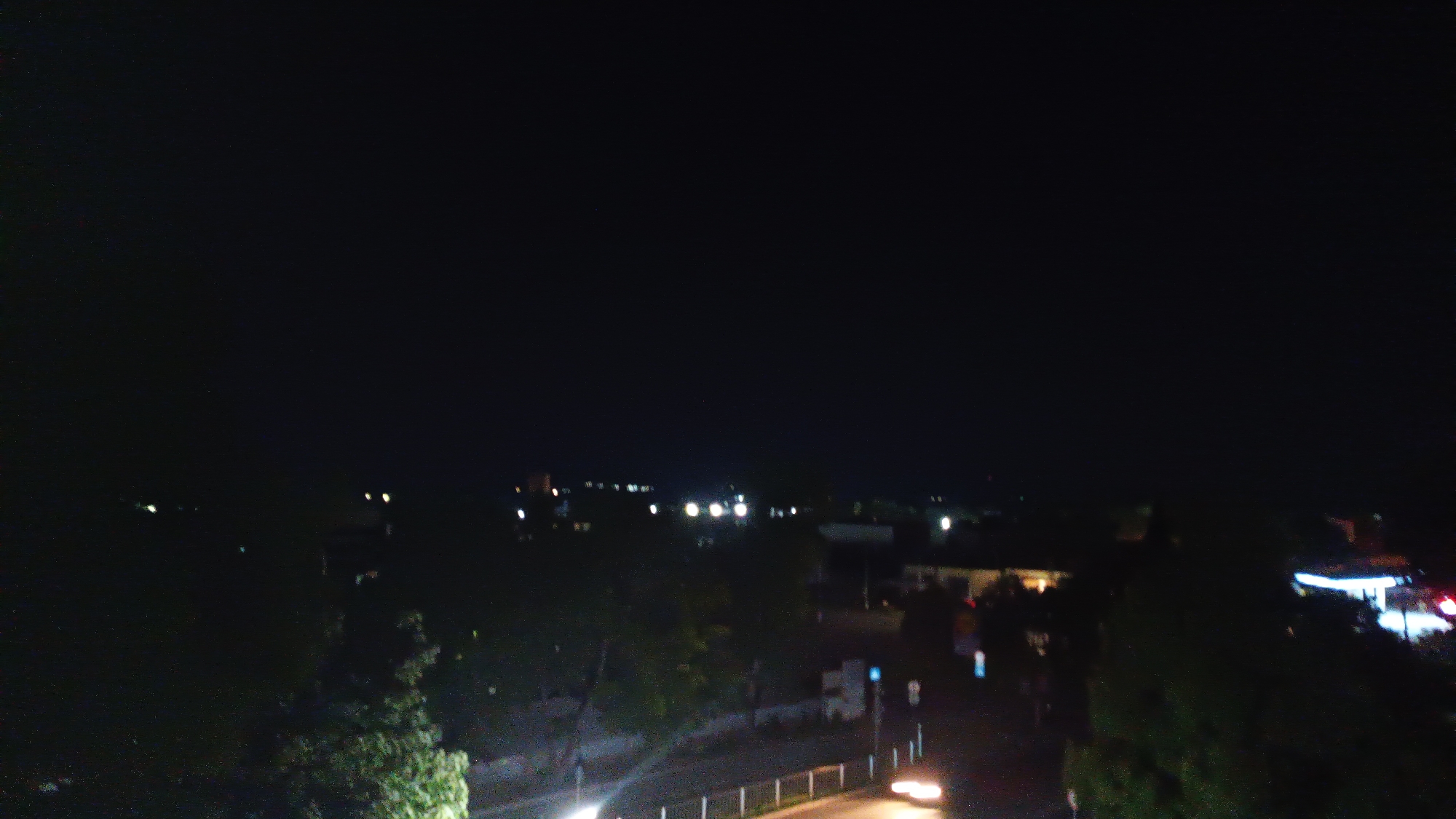 At night, the scene captures a cityscape featuring streetlights, buildings, and trees. The overall environment is dimly lit, with pockets of illumination from the scattered light sources. A road winds through the scene, with a vehicle’s headlights visible.