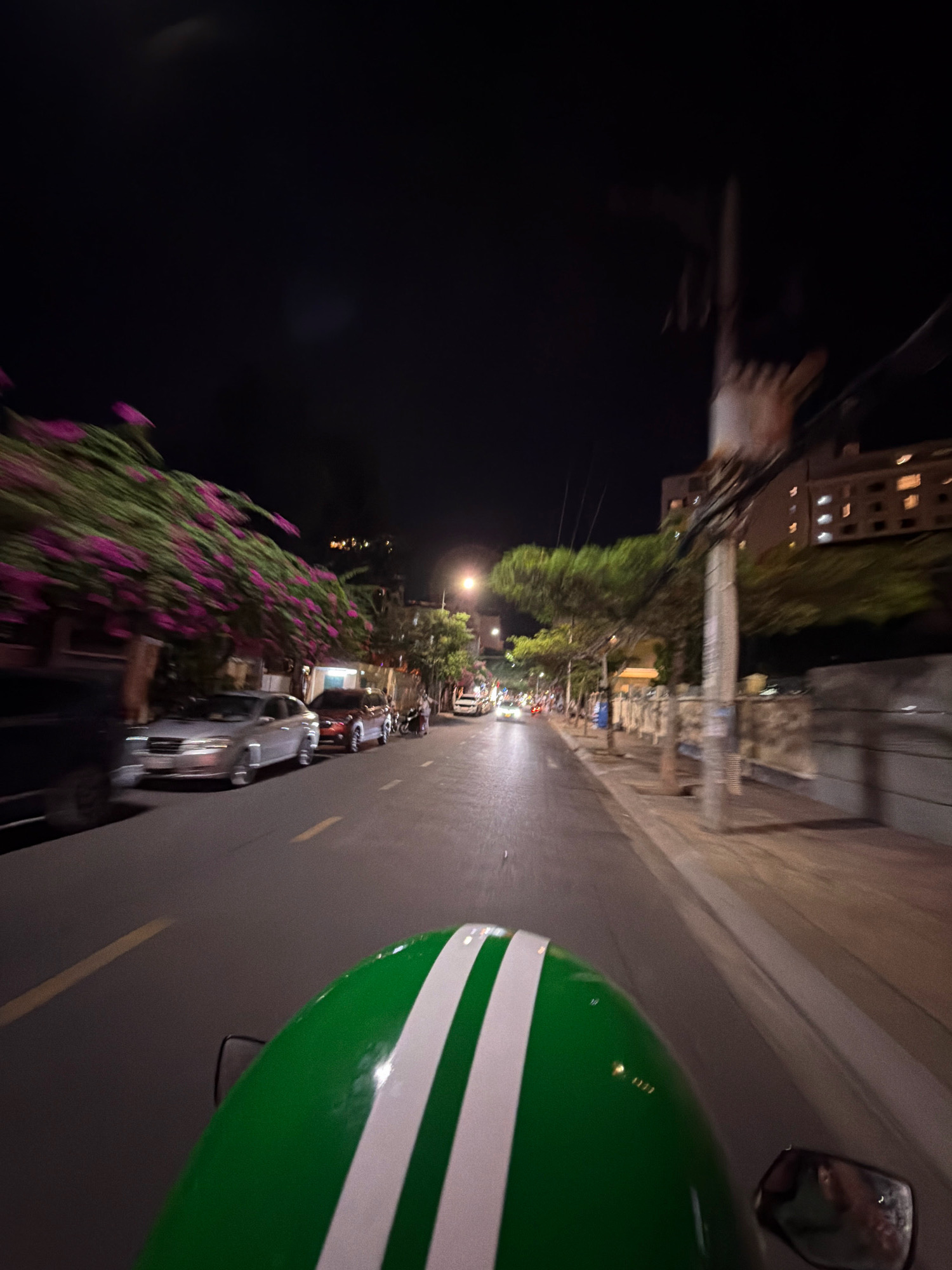 The image captures a point-of-view shot from a moving vehicle, likely a motorcycle, at night in Nha Trang, Vietnam. In the foreground, the rounded, green helmet of the rider with two white diagonal stripes is visible. The road ahead is asphalt, marked with yellow lines. On the left side of the road, a line of parked cars is visible, including a silver sedan. Further down the street, trees with vibrant pink flowers are in bloom, their branches extending over the sidewalk. The right side of the road features a sidewalk, stone walls, and buildings, with some windows illuminated. A utility pole with numerous wires is prominent on the right. The background is dark, indicating nighttime, with distant lights suggesting urban development. The motion blur indicates the vehicle is in transit, likely on a street within Nha Trang.