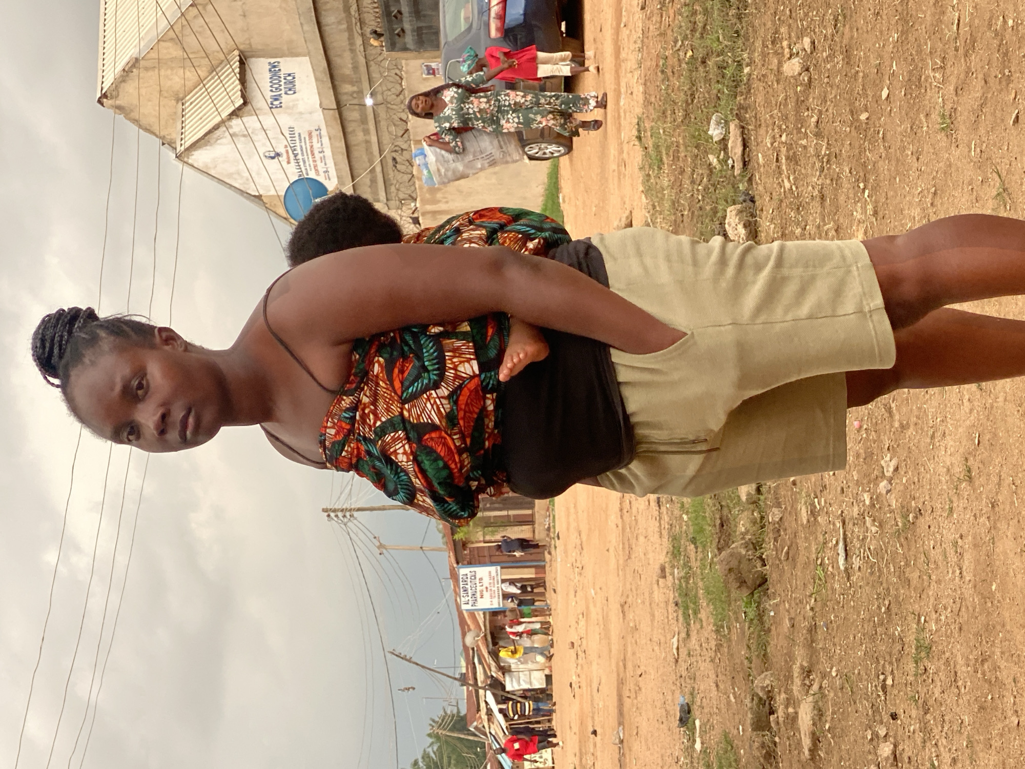 A woman in the foreground, carrying a baby on her back, stands in a dusty, outdoor environment. She is wearing a brightly patterned wrap around her upper body, a black tank top, and light-colored shorts with a drawstring. Her hair is styled in braids and pulled up in a bun. The baby's foot is visible.
Behind her, the scene depicts a street in Sabon Tasha, Nigeria. A building with the text "ECWA GOODNEWS CHURCH" is visible in the background. To the left, a row of shops with signage, including "AL SAMPAROA PHARMACEUTICALS NIG. LTD.", lines the street. Several people are seen walking and standing in the middle ground. A woman in a floral-patterned jumpsuit is carrying a large sack over her shoulder, and a child in a red shirt and green headscarf walks beside her. A blue car is parked near them. The ground is a mix of dirt and scattered debris. The sky is overcast.