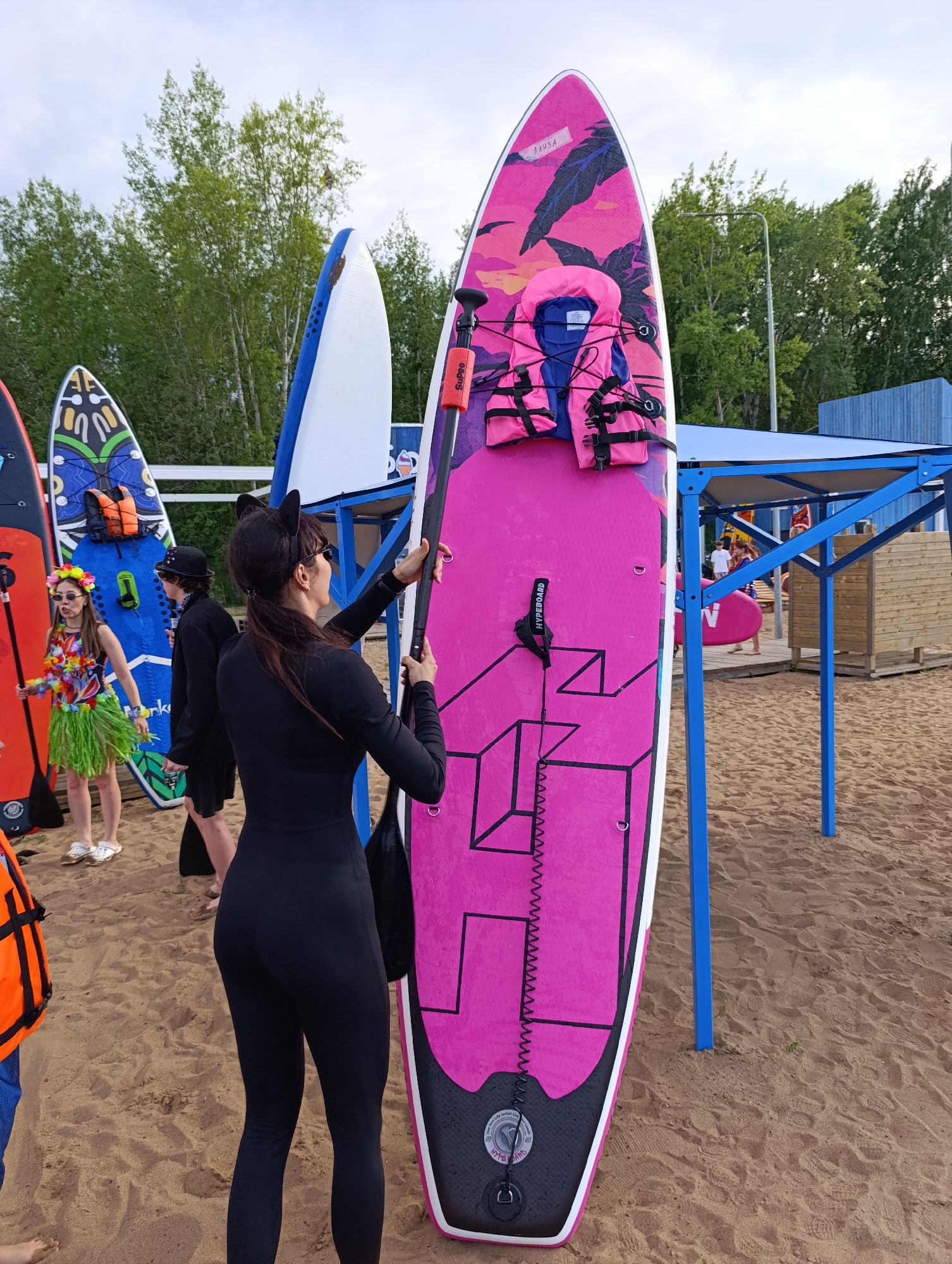 Two women and a person in a black suit stand on a sandy beach near a body of water, with several paddleboards lined up. The woman in the foreground, wearing a black wetsuit and cat ears, holds a paddle. She is looking to the right, towards another woman dressed in a colorful Hawaiian outfit with a grass skirt. Behind them, more paddleboards and a blue structure with a canopy are visible. A third person in a black suit and hat stands between the two women. The paddleboards are varied in color and design, with one large pink board in the foreground featuring a tropical pattern and a life vest attached. A pink paddleboard is seen being carried by a person under the blue canopy in the background. The weather appears overcast, suggesting it is either early morning or late afternoon. The scene suggests a recreational setting, possibly related to paddleboarding or a beach event. The visible text on the pink paddleboard reads "NUUSA" and "HYBEBOARD".