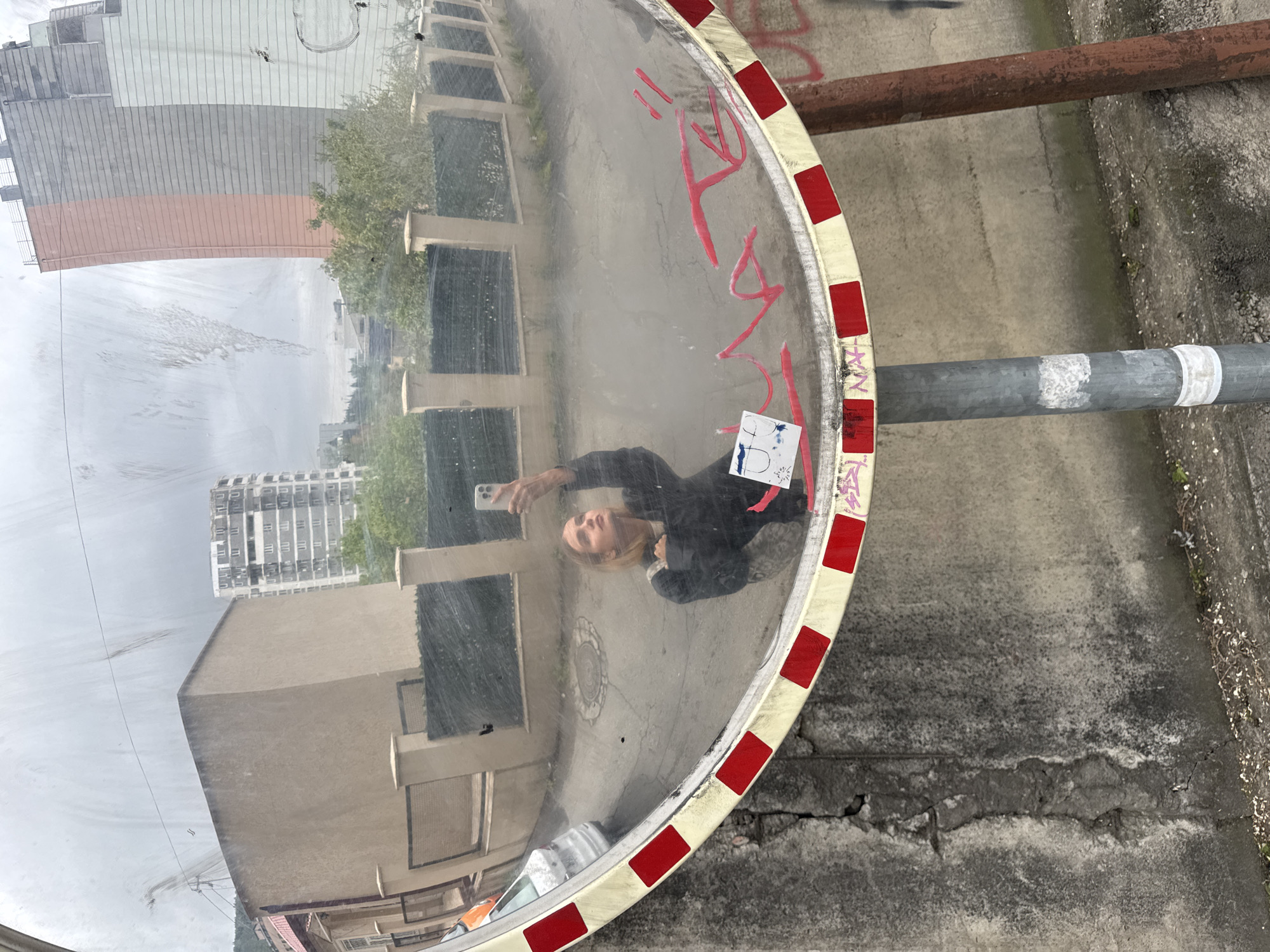 The image depicts a convex traffic mirror reflecting a street scene, with a person taking a selfie visible in the mirror's reflection. The mirror has a white and red checkered border and is mounted on a metal pole. Graffiti is present on the mirror's surface.
Reflected in the mirror are buildings of varying heights and architectural styles, suggesting an urban environment. The street pavement and a segment of a brown pipe are also visible in the reflection. The person taking the selfie has blonde hair and is wearing a dark jacket.
Based on visual analysis, the location is likely Tbilisi, Georgia, considering the architectural style of the buildings in the background. Further contextual information or metadata from the image source would be needed for definitive confirmation.