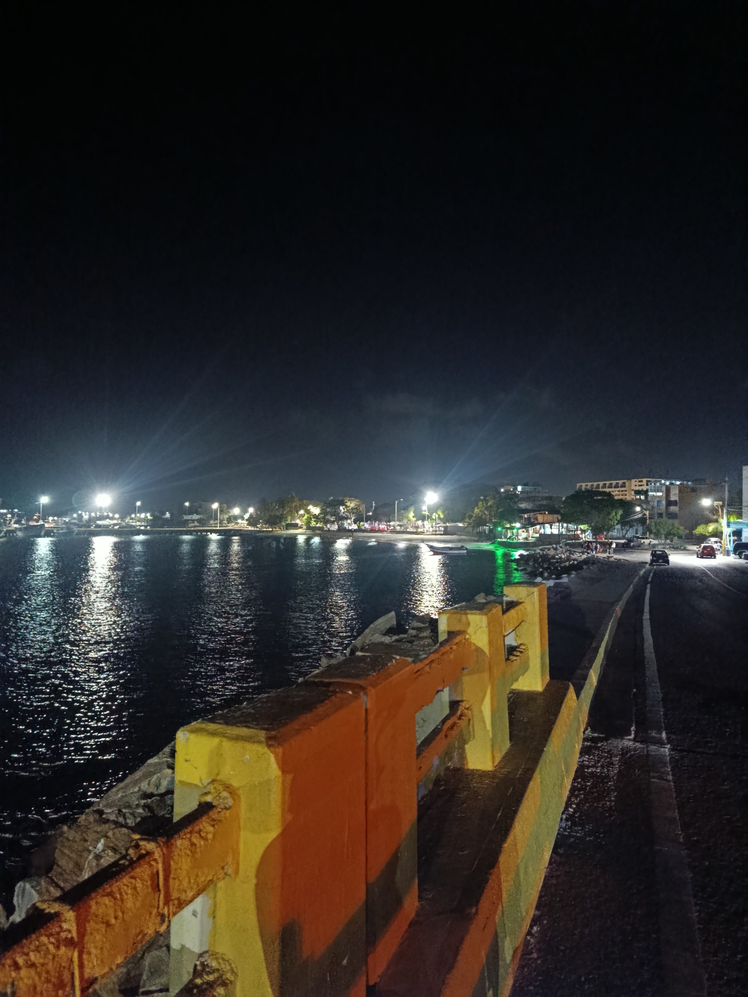The image depicts a nighttime scene in Juan Griego, Venezuela. The view is from a waterfront walkway or road, with a brightly painted orange and yellow barrier in the foreground. To the left, the dark water of the bay reflects the lights along the shoreline. The shoreline is dotted with streetlights and buildings, some illuminated with green light. A few cars are visible on a road along the shore. Above the shoreline, the night sky is dark. To the right of the barrier, a road with a white line marking runs along the edge of the water.