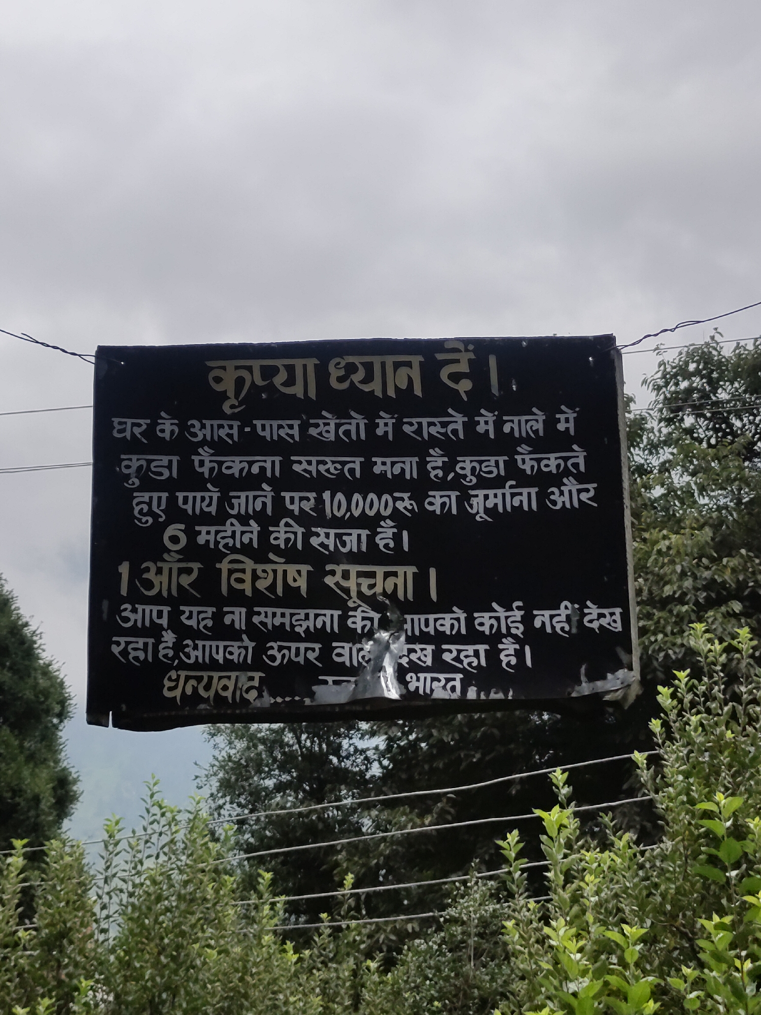 A black, rectangular sign with white Hindi text is mounted on a pole against a cloudy sky. The sign reads: "कृपया ध्यान दें । घर के आस-पास खेतों में रास्ते में नाले में कूड़ा फेंकना सख़्त मना है, कूड़ा फेंकते हुए पाये जाने पर 10,000 रुपये का जुर्माना और 6 महीने की सज़ा है। 1 और विशेष सूचना । आप यह ना समझे की आपको कोई नहीं देख रहा है, आपको ऊपर वाले देख रहा है। धन्यवाद ....... भारत ।" This translates to: "Please pay attention. It is strictly forbidden to throw garbage in the fields, on the paths, and in the drains near the houses. If found throwing garbage, there is a fine of Rs. 10,000 and a sentence of 6 months. 1 more special notice. Do not think that no one is watching you, God is watching you. Thank you....... India." The sign appears weathered, with some peeling and water damage, particularly along the bottom edge. Lush green trees and foliage are visible in the foreground and along the bottom of the frame, suggesting an outdoor, possibly rural or park-like setting. Electrical wires run horizontally across the upper part of the image, above the sign. The overall scene suggests a public notice aimed at maintaining cleanliness and order.