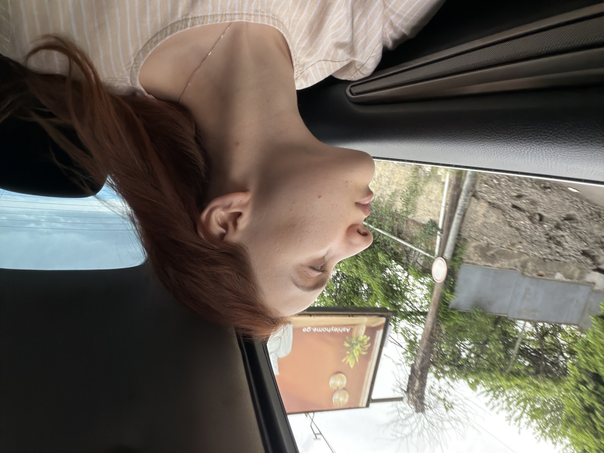 A young, light-skinned woman with auburn hair is partially visible inside a car, likely as a passenger. She is wearing a striped shirt and a delicate necklace. The car's window shows an outdoor scene, revealing an advertisement board for "ashleyhome.ge" indicating the city is likely Tbilisi, Georgia. The background features trees and a concrete building under daylight conditions. The image is captured from inside the car, focusing on the woman and the external environment visible through the window. The perspective is slightly tilted.