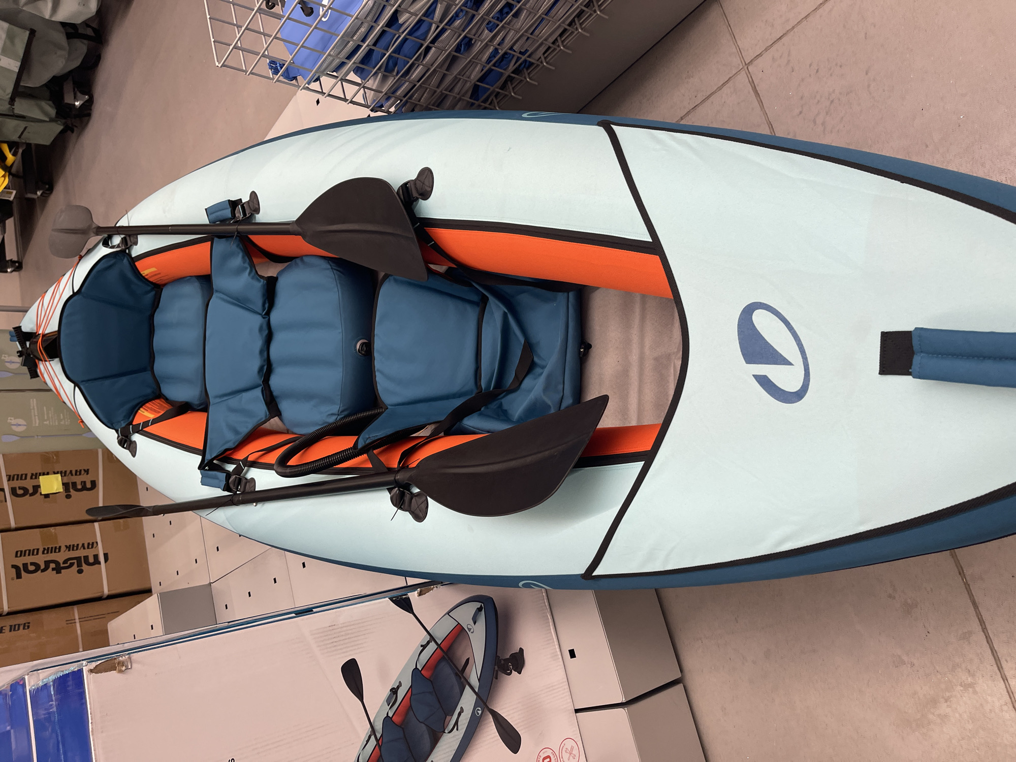 The image shows a light blue and orange inflatable kayak with two black paddles. The kayak has blue life vests inside. The kayak is sitting on the floor of a retail store, surrounded by boxes of the same kayak. The text on the boxes reads "Mistral Kayak Air Duo." The location is likely a store in Sofia, Bulgaria.