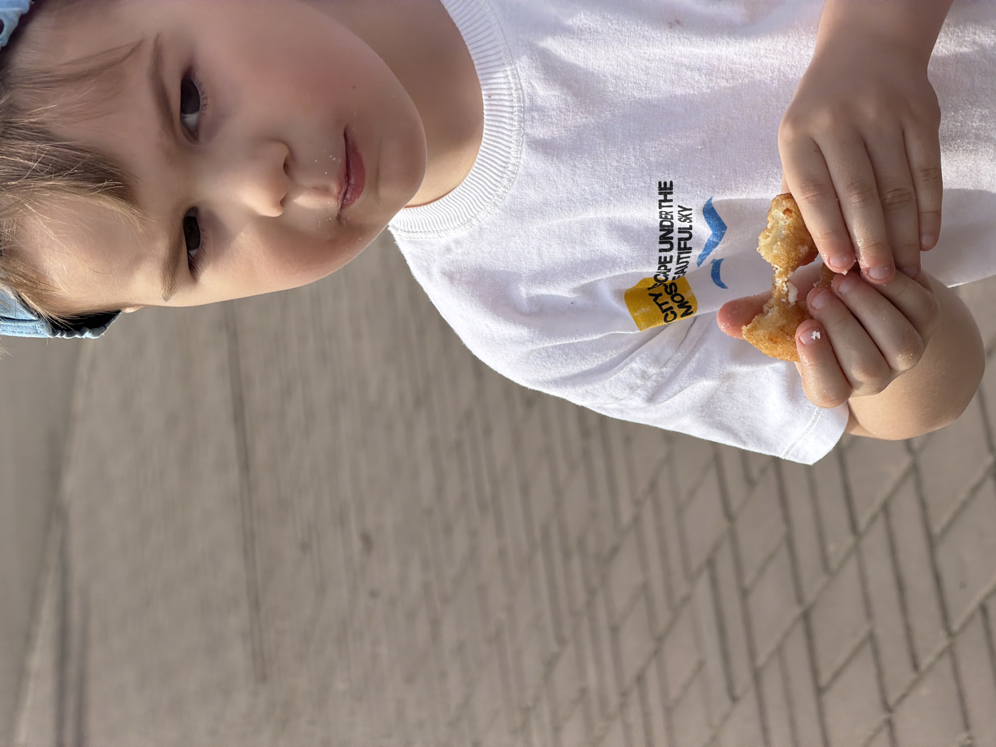 A young child with light hair and a blue cap stands outdoors, holding a partially eaten fried dough ball in their hands. The child is wearing a white t-shirt with the words "CITY MOSCOW ESCAPE UNDER THE BEAUTIFUL SKY" printed on it. The ground is paved with light brown bricks. This photo was taken in Krasnoyarsk, Russia.