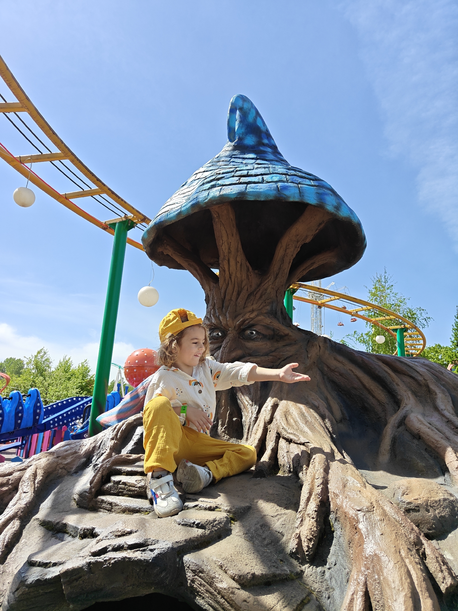 A young child with light brown curly hair, wearing a yellow hat, a white shirt with rainbow designs, and yellow pants, is sitting on a rocky, tree-like structure with a blue mushroom-shaped roof. The structure has a face carved into the trunk. The child is extending their right arm with an open palm. In the background, there is a roller coaster track, other amusement park rides, and trees. The sky is blue with some clouds. This image was taken in Khimki, Russia.